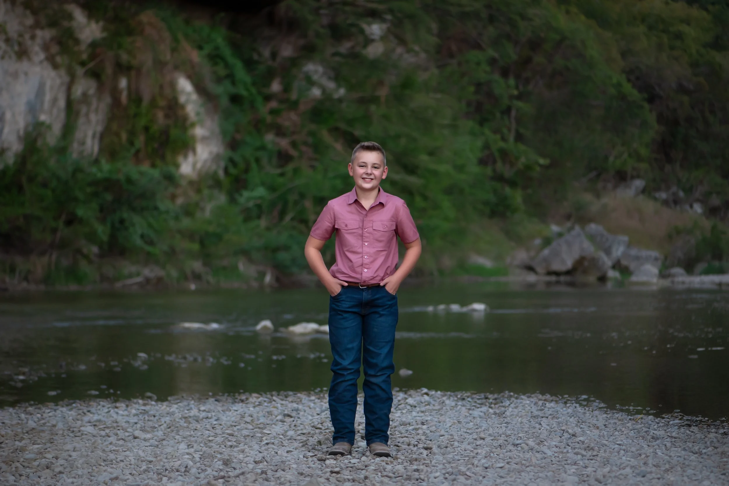 A young boy with short hair, wearing a pink button-up shirt and blue jeans, standing on a rocky riverbank with hands in his pockets, smiling at the camera, with a river and green trees in the background.