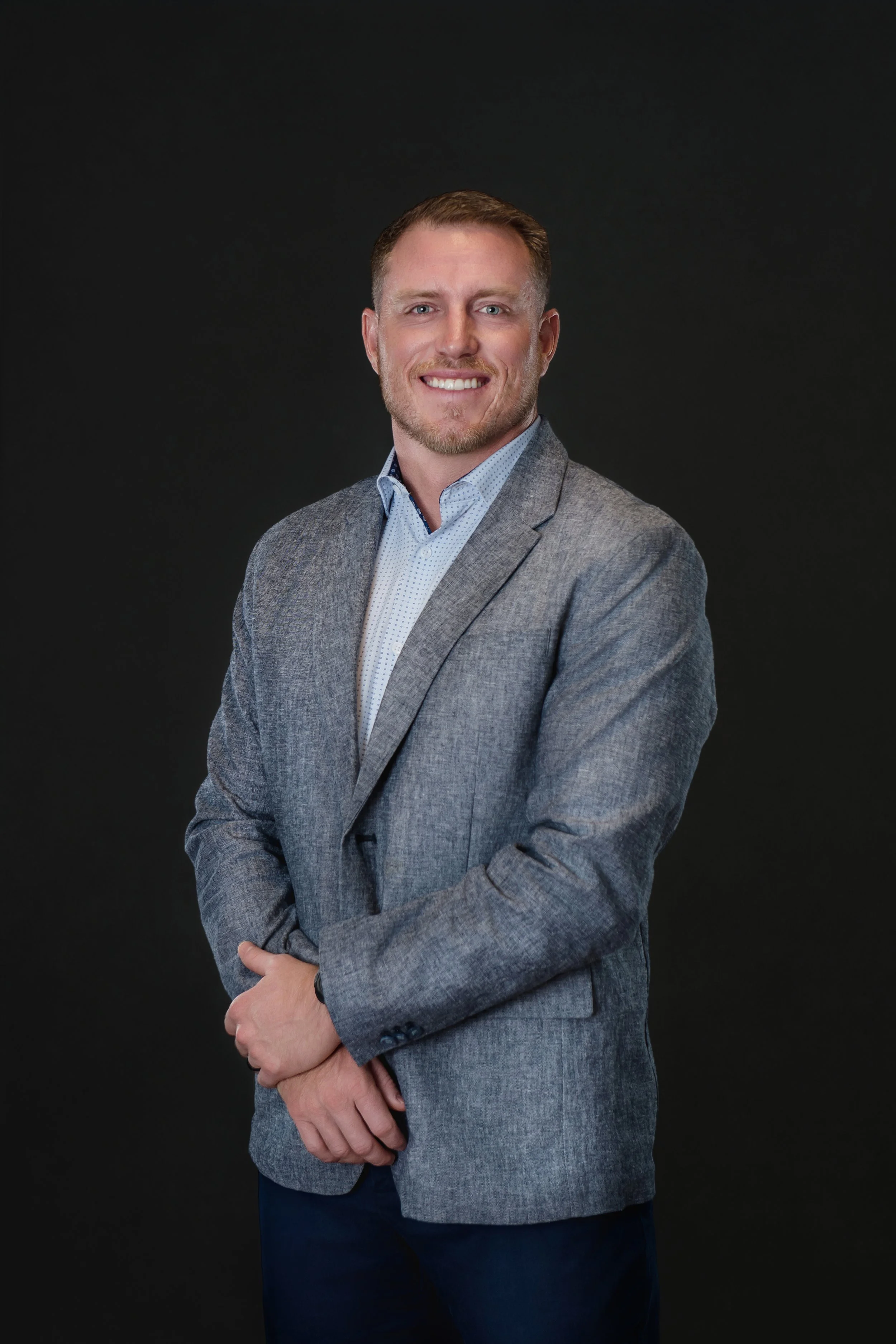 Portrait of a man wearing a gray suit jacket and light blue shirt, standing against a dark background, smiling with arms crossed.