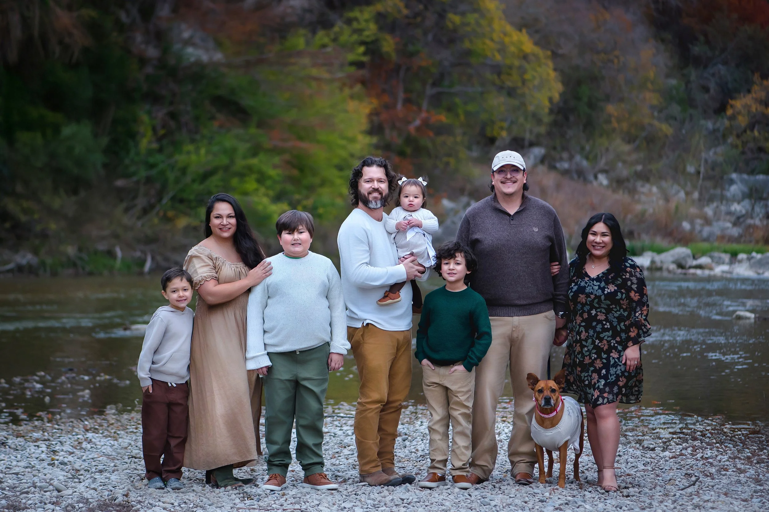 A family of nine, including six children and two adults, standing on a rocky riverbank with trees and a river in the background during fall.