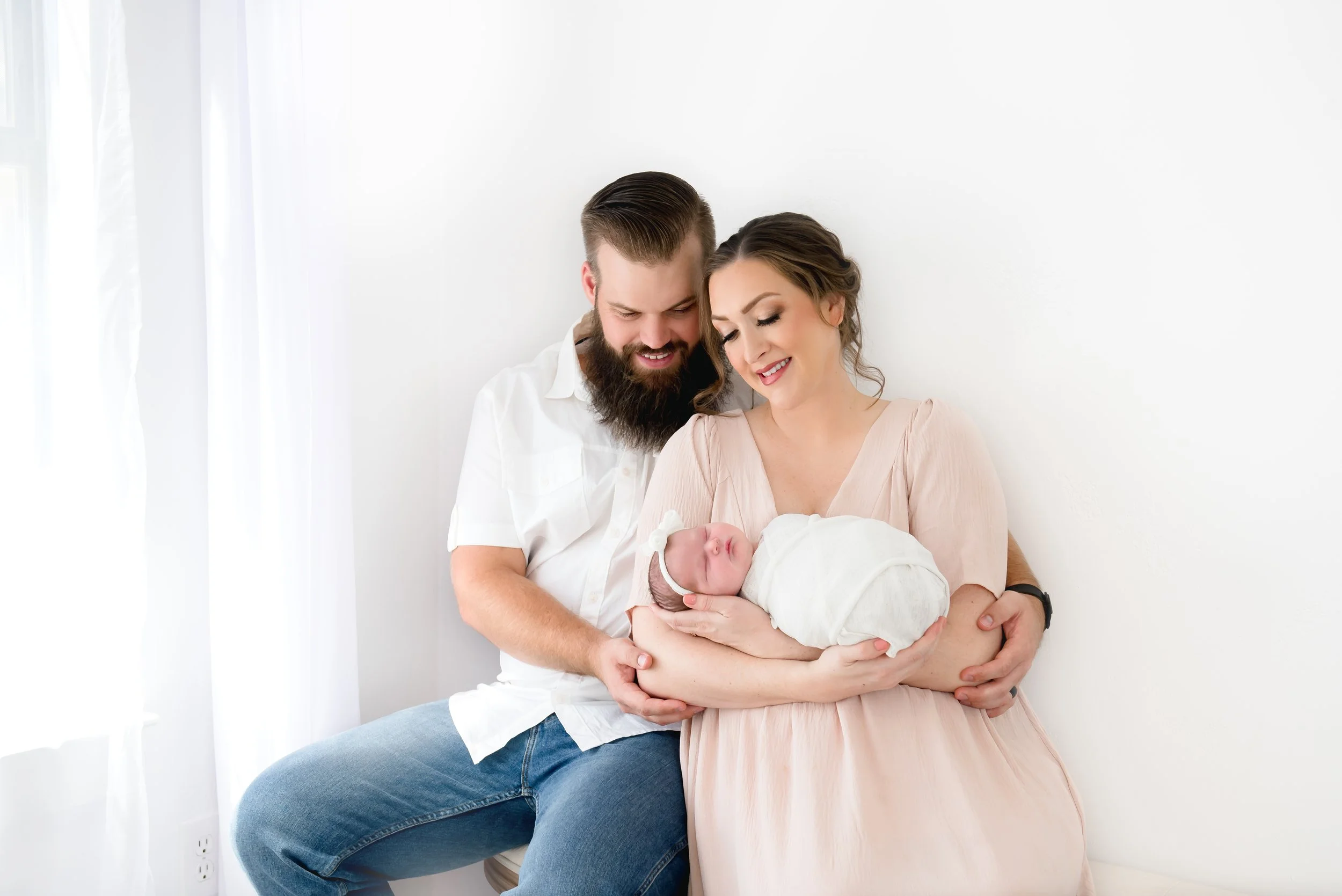 A happy family holding a newborn baby near a white wall with white curtains.