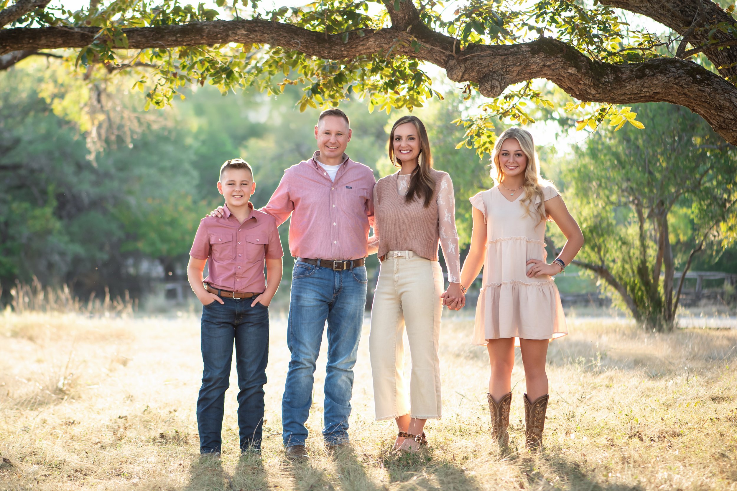 A family of four standing outdoors under a tree, holding hands, smiling, with sunlight filtering through the leaves, in a grassy area surrounded by green trees.