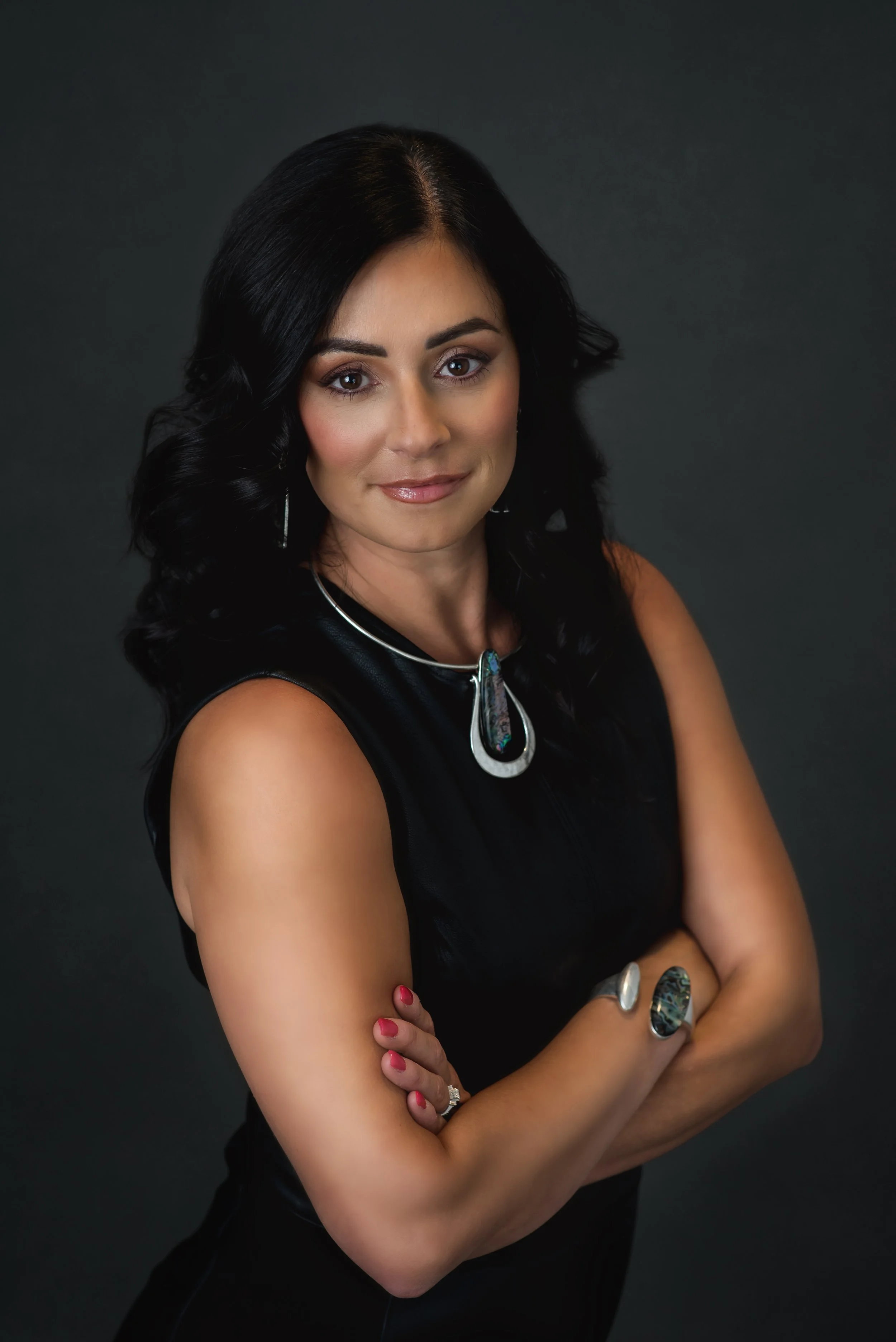 A woman with black hair wearing a black sleeveless top and jewelry, looking confident with arms crossed against a dark background.
