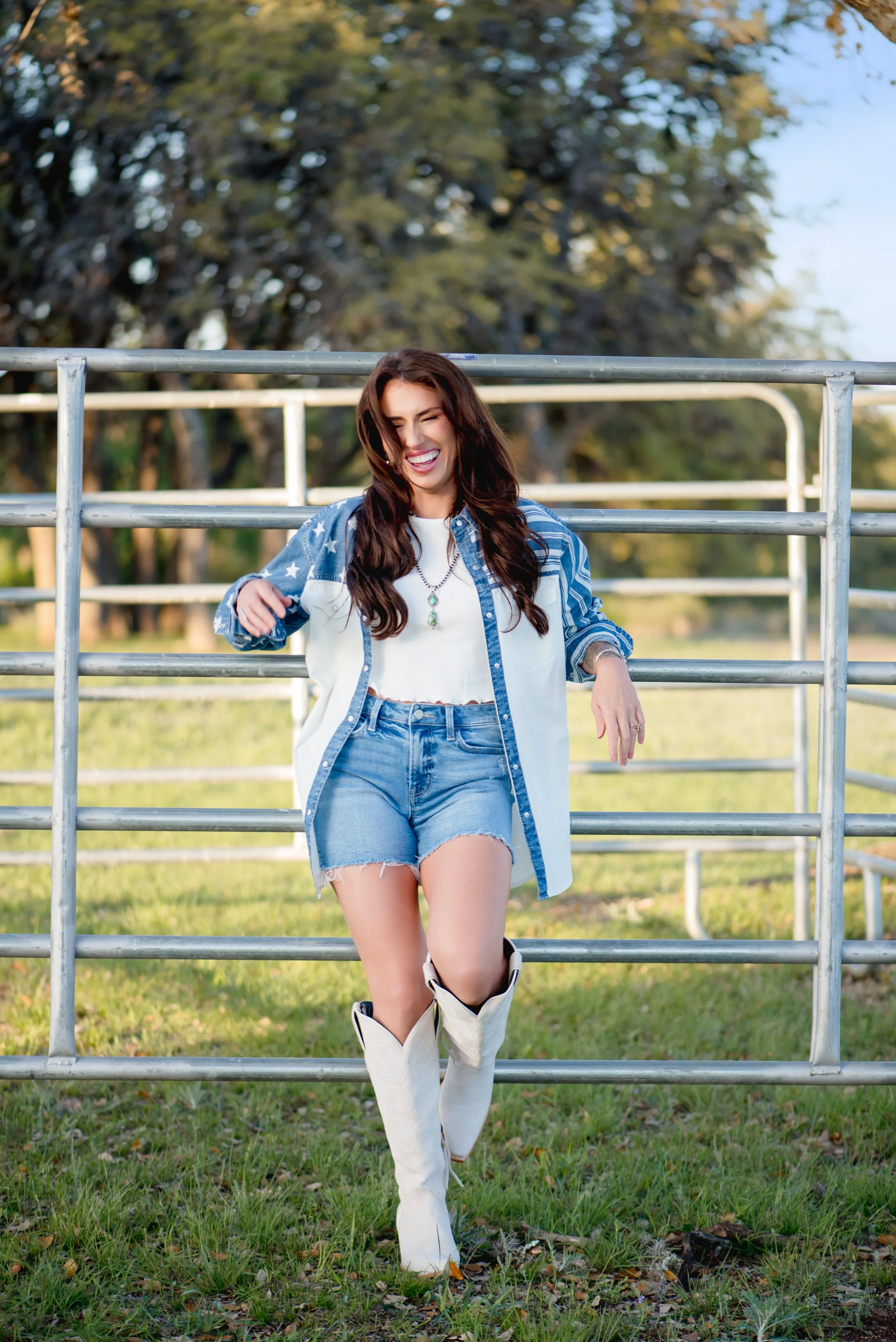 A woman with long brown hair wearing a white top, denim shorts, a button-up shirt with star patterns, and white cowboy boots, smiling and leaning against a metal fence outdoors in a grassy field with trees in the background.