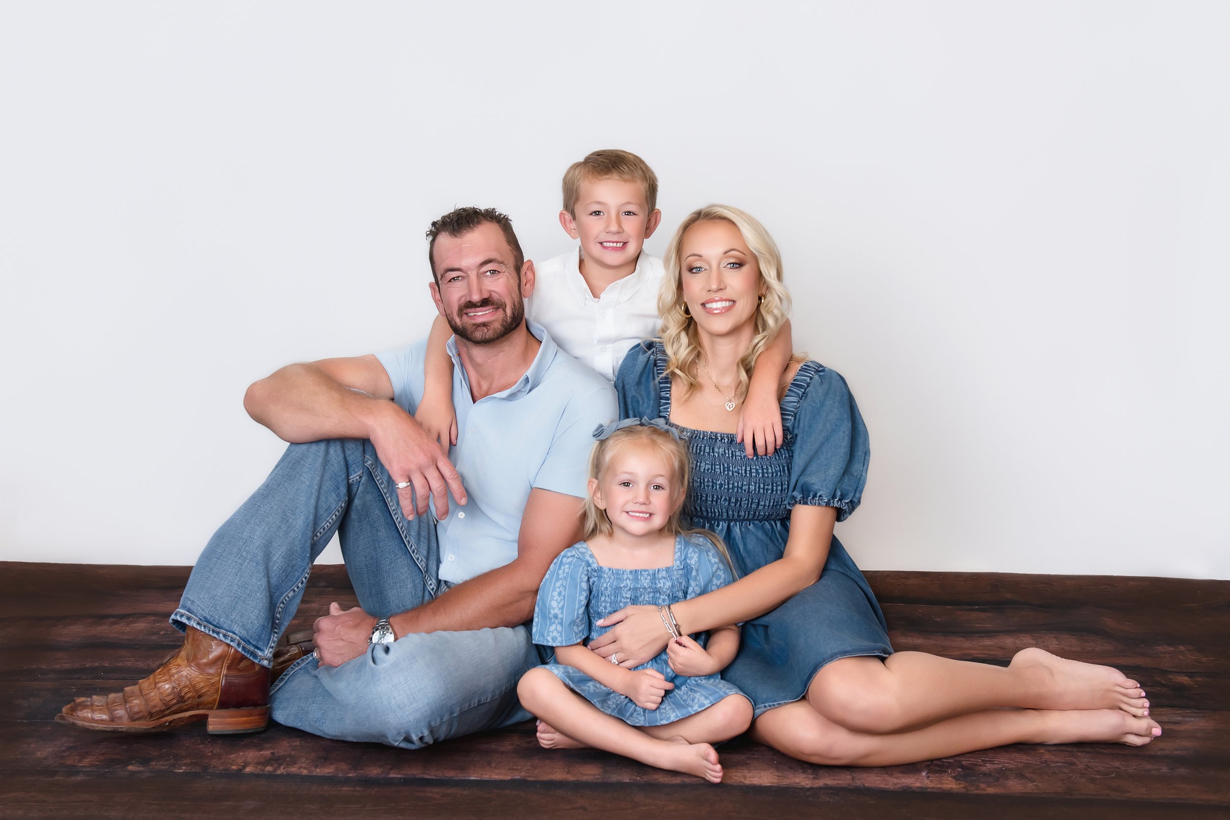 A family of five posing together on a wooden floor against a plain white background. The family includes a father, mother, two daughters, and a son. The father is sitting on the floor with his legs crossed, wearing a light blue shirt and jeans. The m