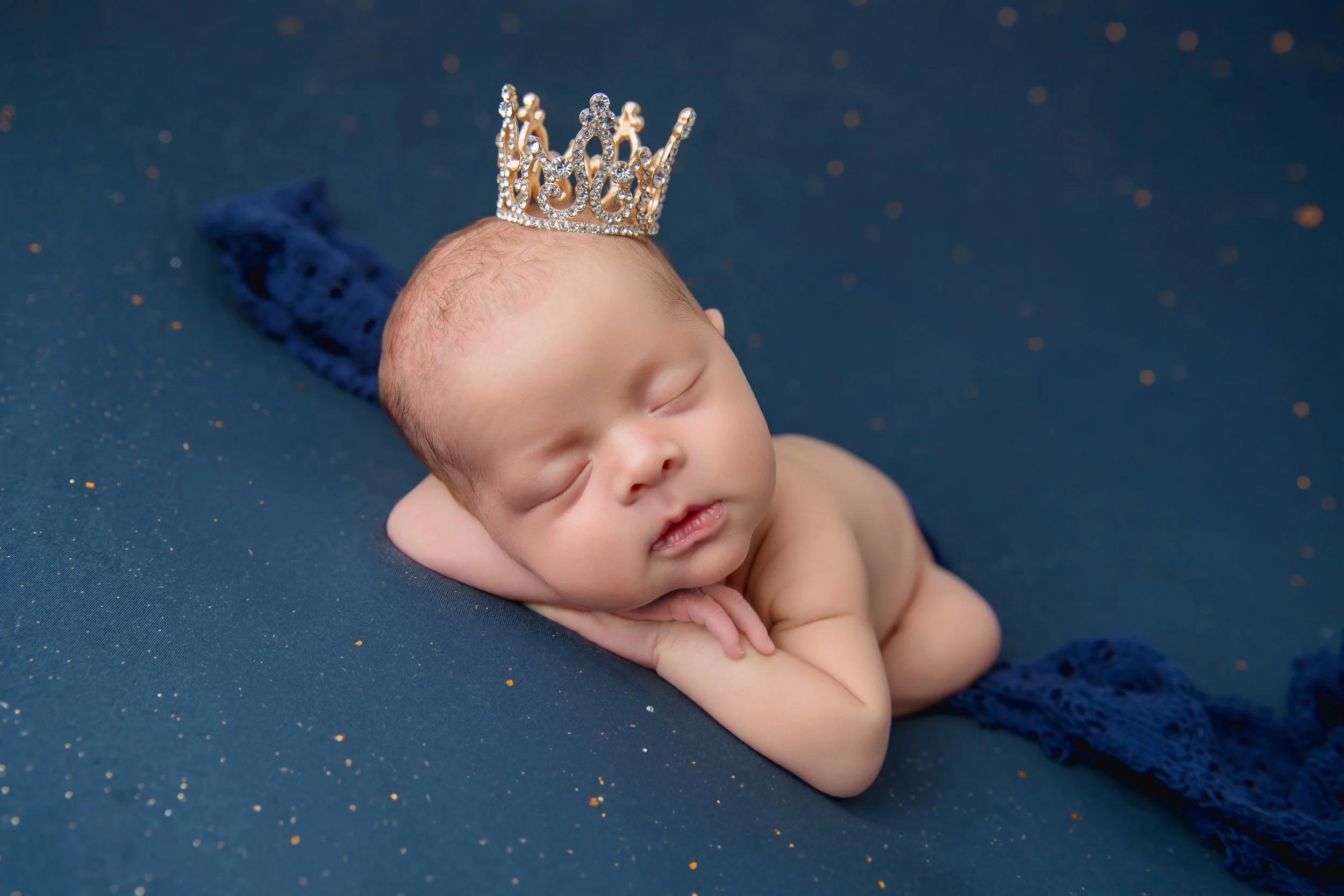 Editorial newborn portrait of a sleeping baby wearing a jeweled crown, posed on deep blue fabric with gold accents for a regal heirloom look.