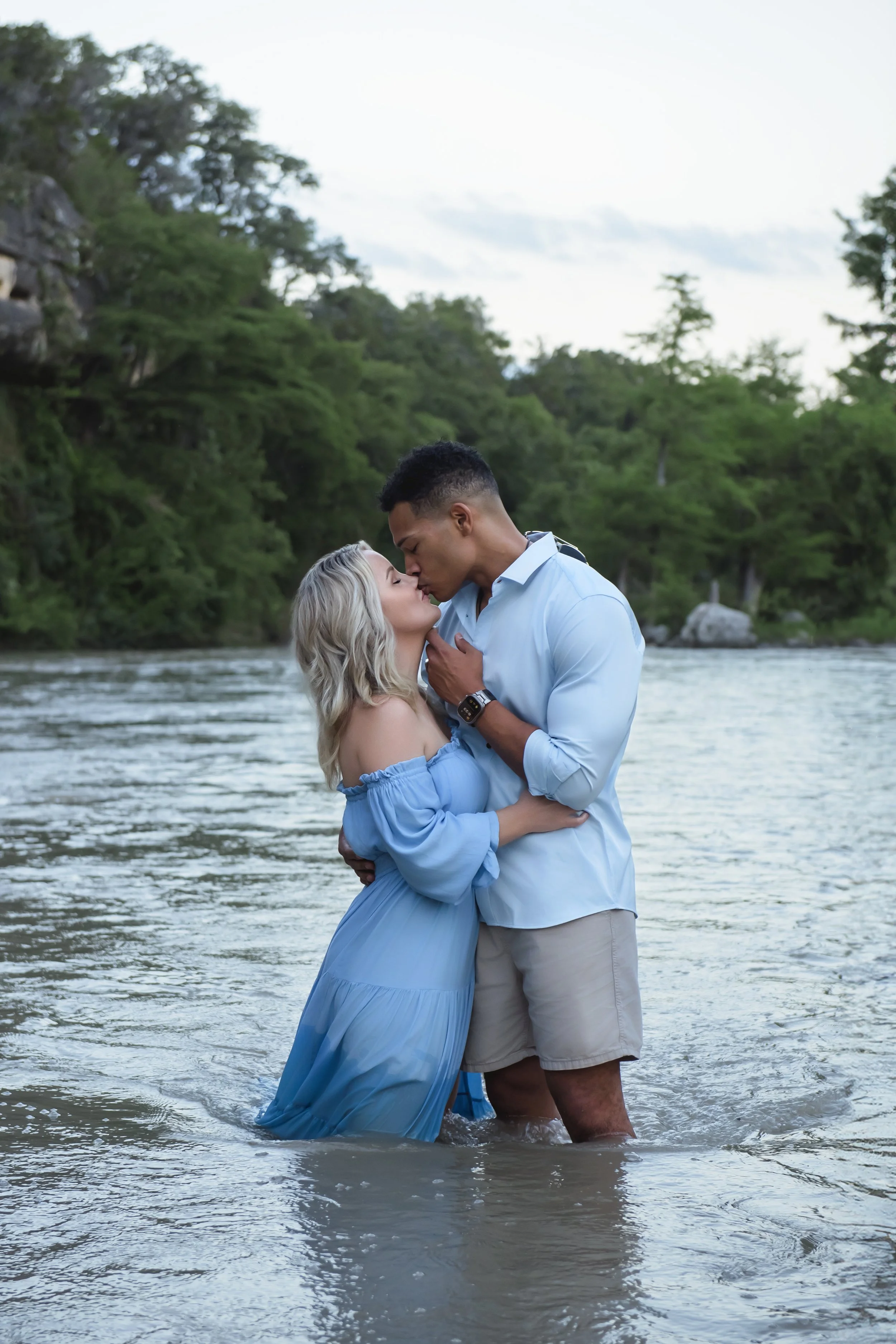 A couple sharing a kiss in a river surrounded by green trees.