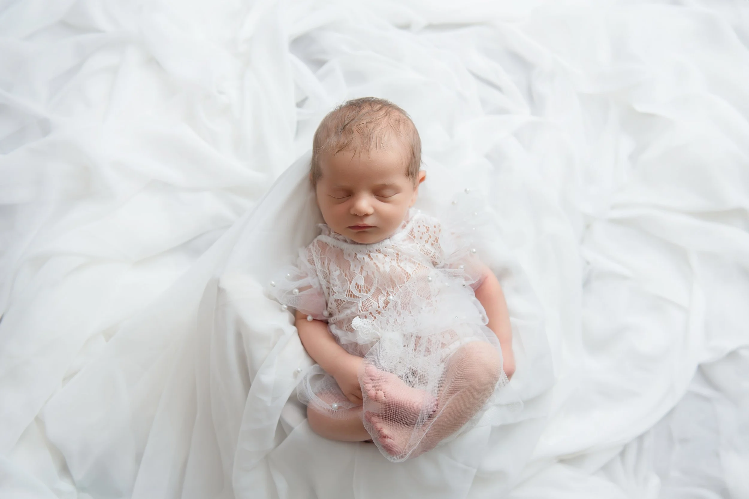 A sleeping newborn baby with light brown hair, dressed in a delicate lace white dress, lying on soft white fabric with pearl embellishments.
