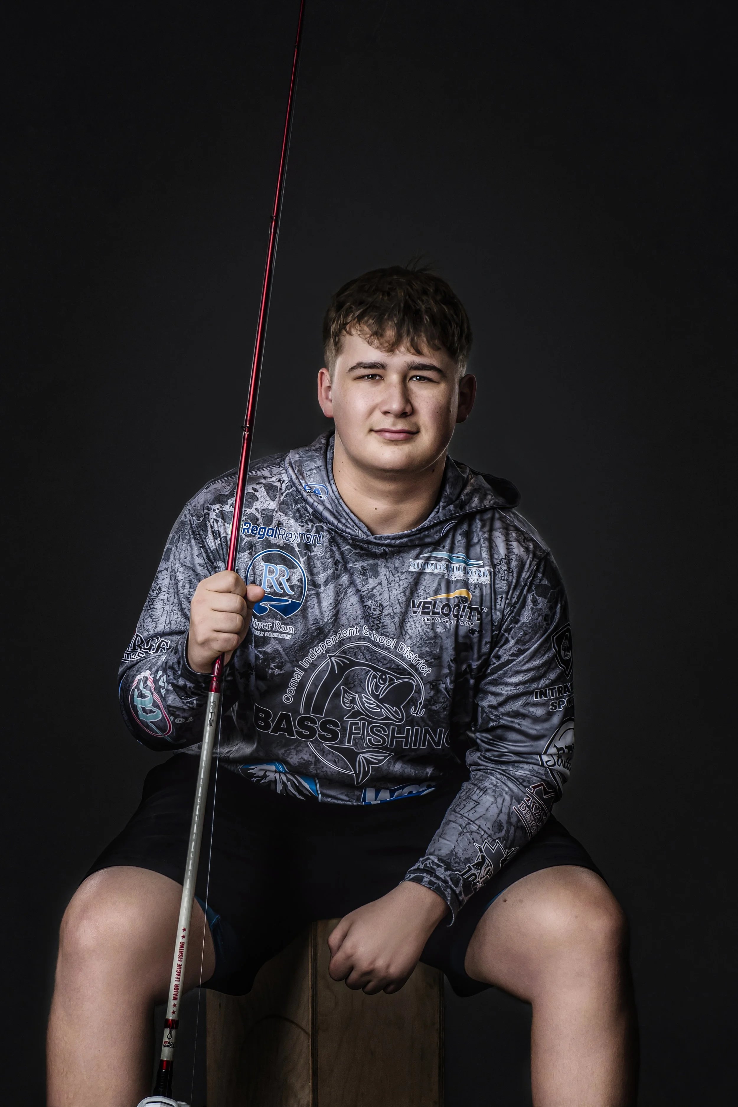 A young man sitting on a wooden stool against a black background, holding a fishing pole, wearing a gray camouflage hoodie with fishing and outdoor logos, and black shorts.
