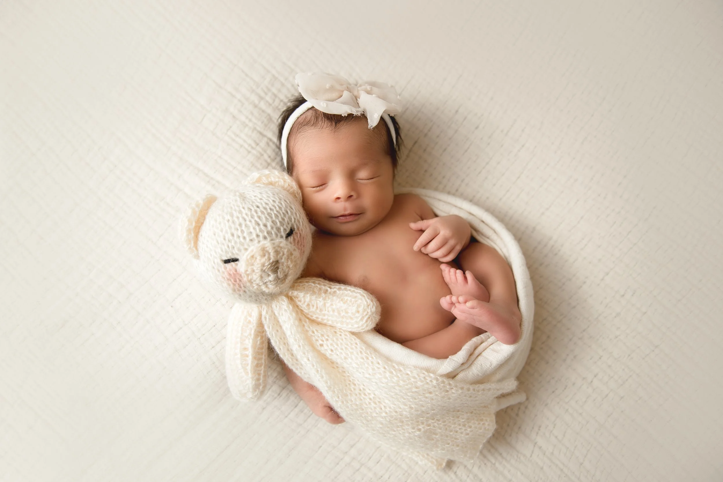 A newborn baby sleeping, wrapped in a cream-colored knitted blanket, with a knitted teddy bear and a white headband with a bow, lying on a cream-colored textured surface.