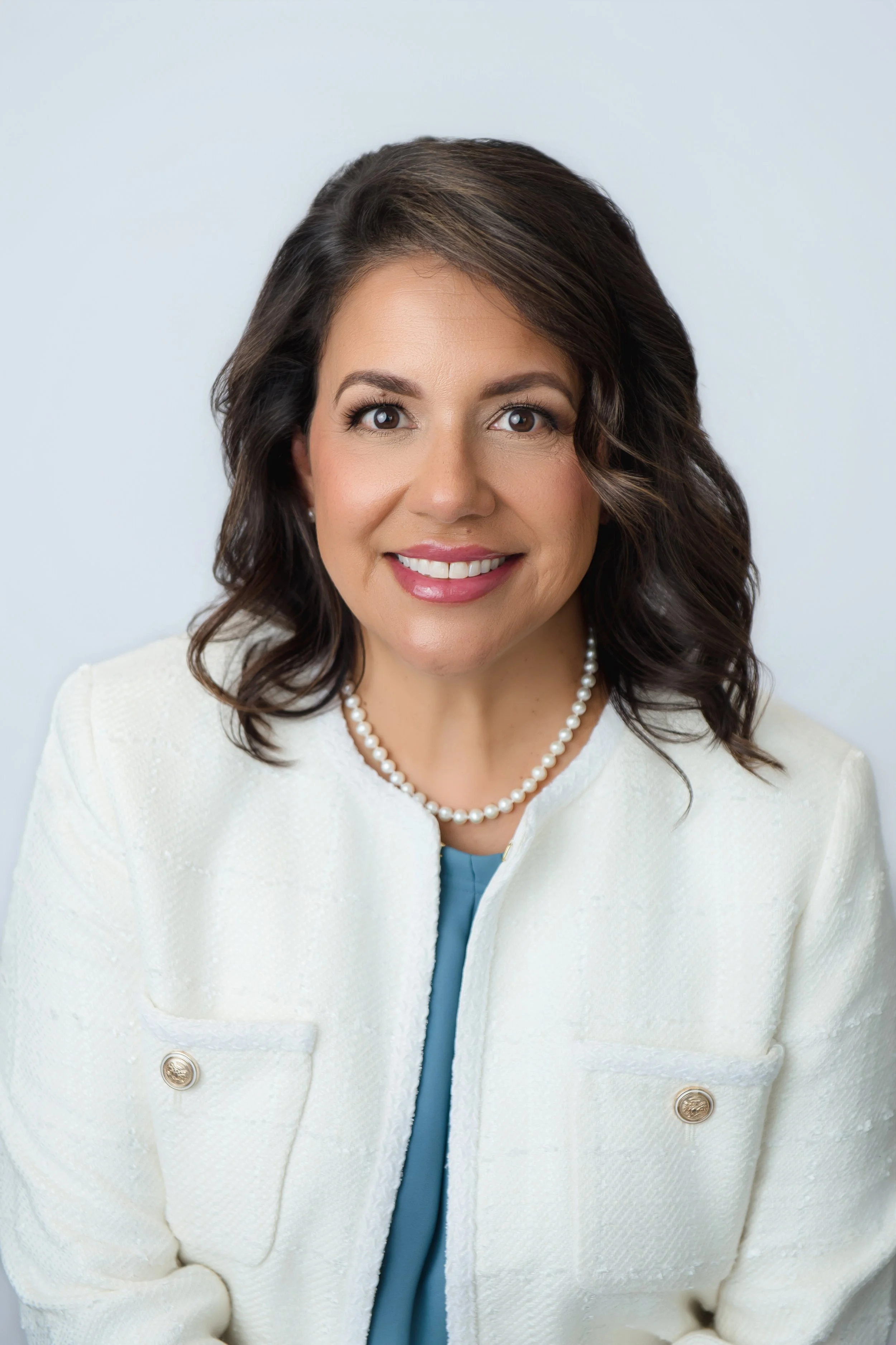 Portrait of a woman with shoulder-length wavy brown hair, wearing a white blazer with pearl buttons, a pearl necklace, and a blue top, smiling at the camera against a plain light background.