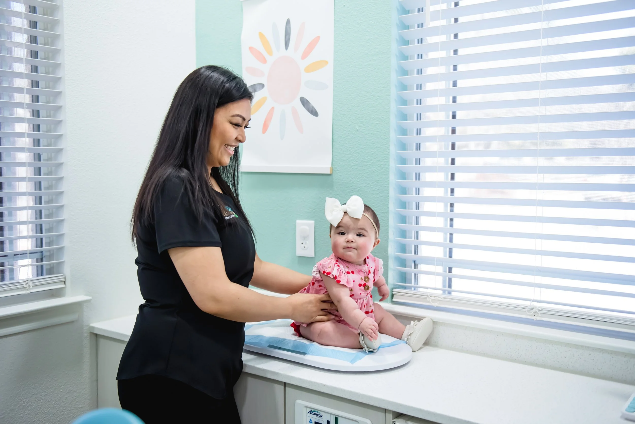 A healthcare professional, wearing a black uniform, is attending to a baby girl sitting on a medical examination table in a bright room. The baby, dressed in a pink outfit with a white bow headband, is looking towards the camera.