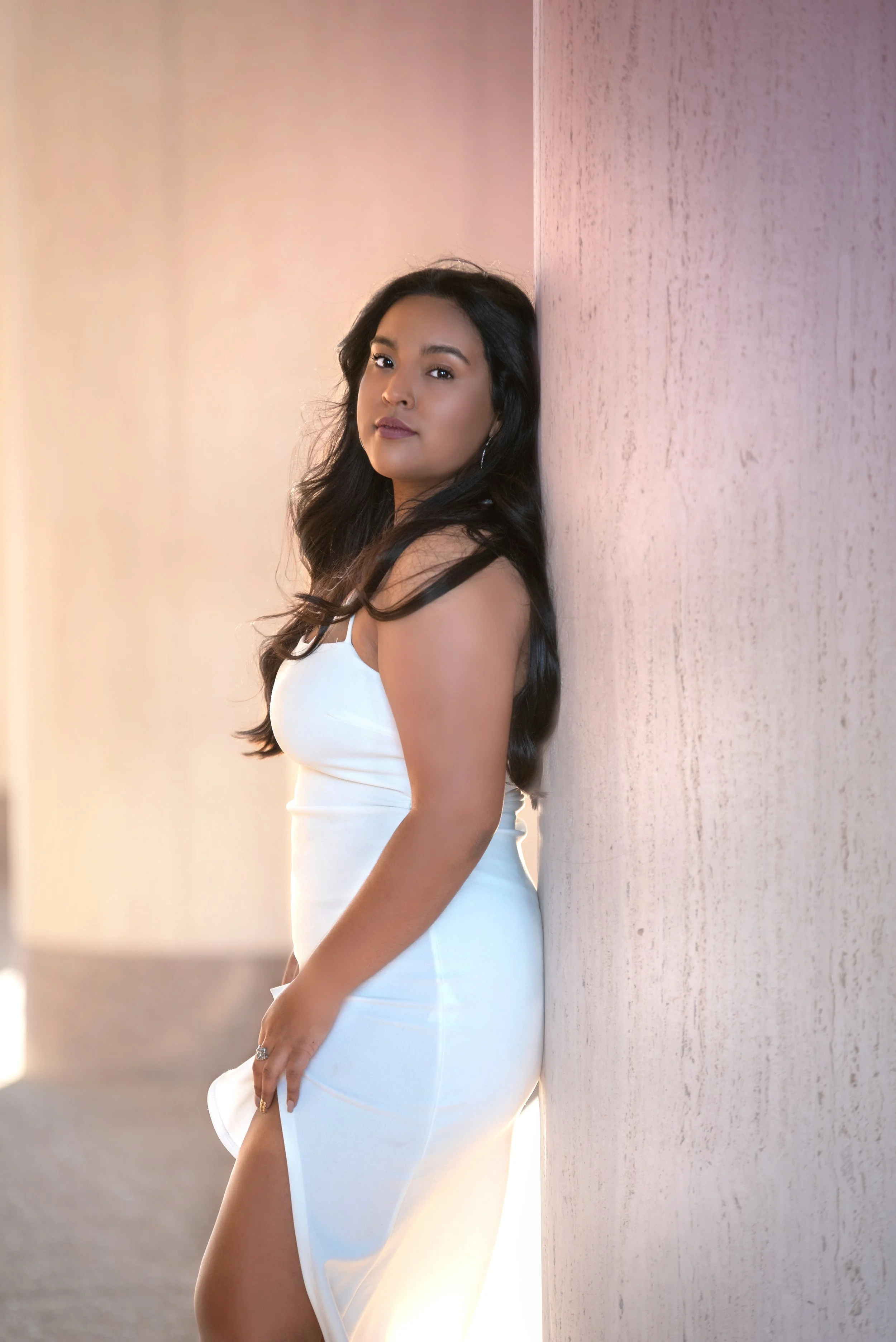 A woman in a white dress leaning against a textured pink wall indoors, with a soft blurred background.