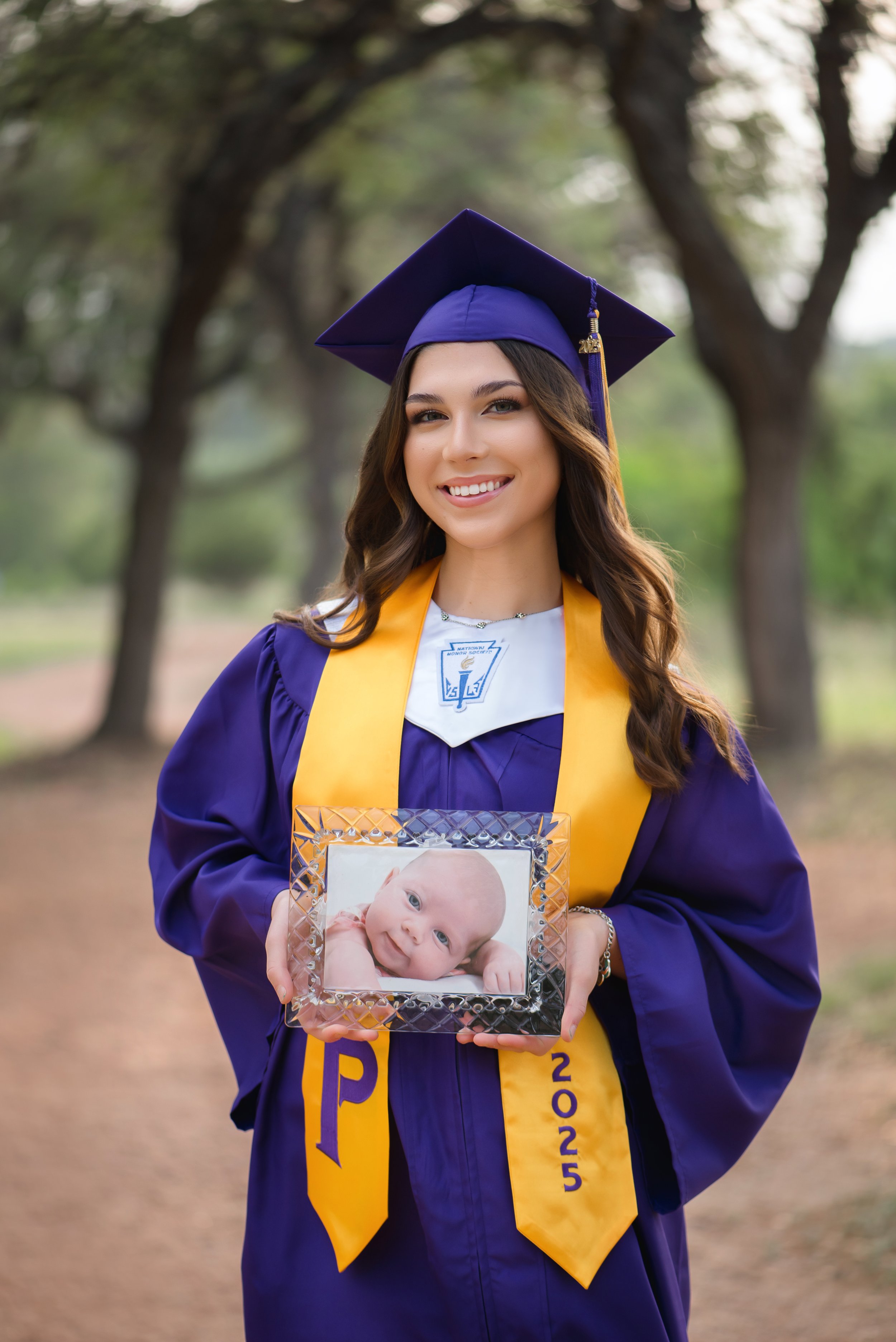 A young woman in a purple graduation cap and gown holding a framed photo of a baby and smiling outdoors.