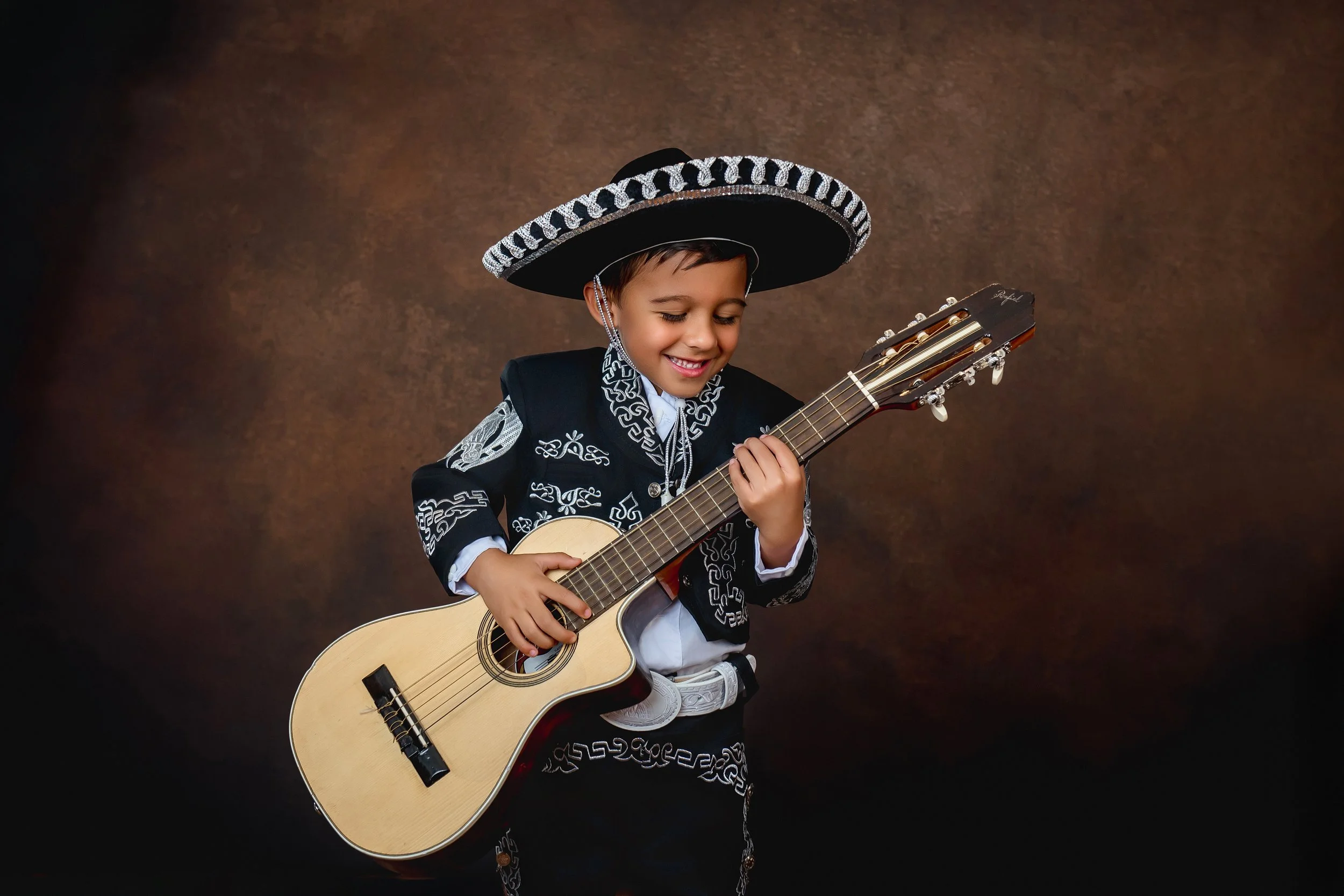 Young boy dressed in traditional Mexican mariachi clothing, playing an acoustic guitar, smiling, against a brown background.