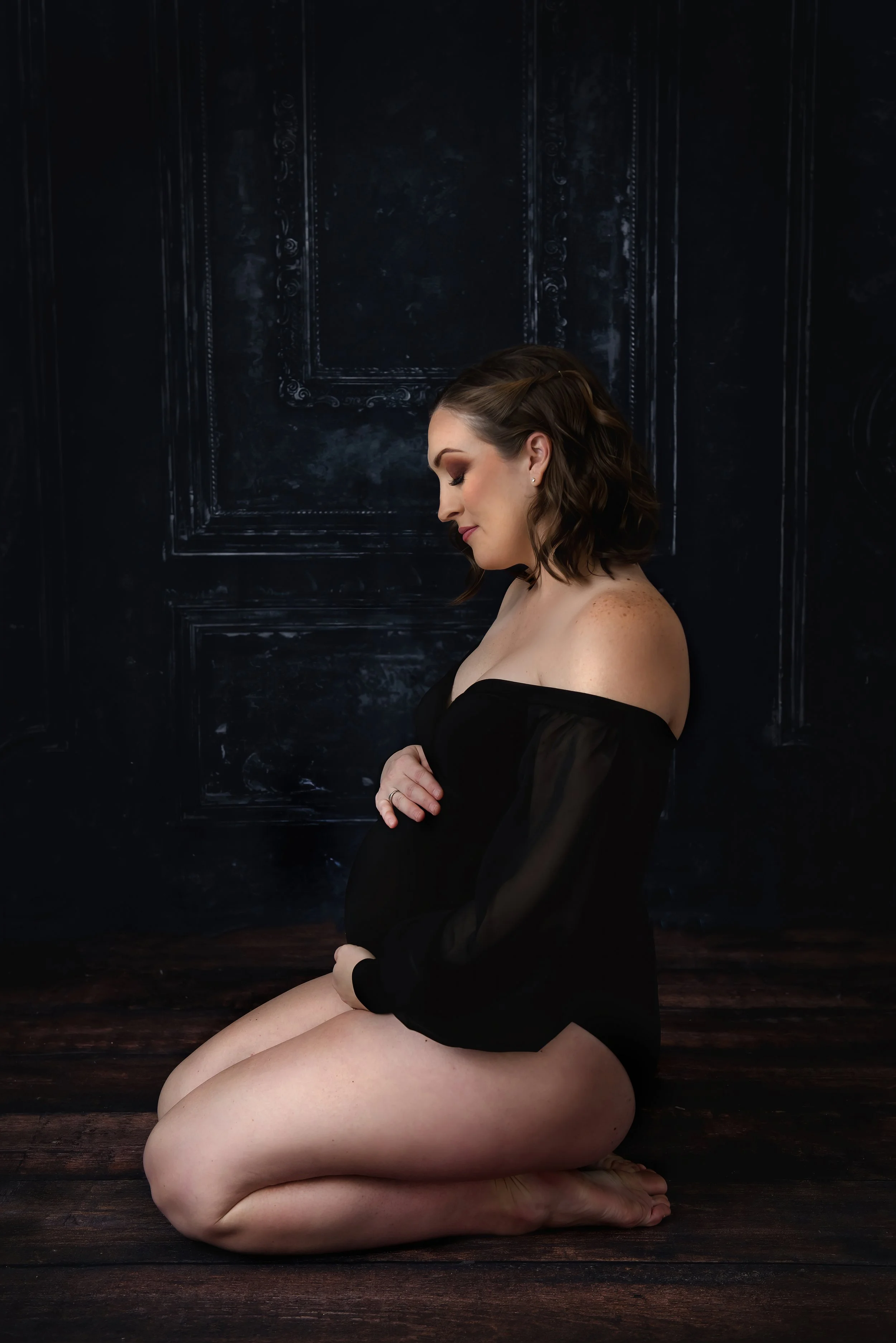 Fine art studio maternity portrait of a pregnant woman kneeling on a dark wooden floor, wearing an off-shoulder black top and resting her hands on her baby bump against a textured backdrop.