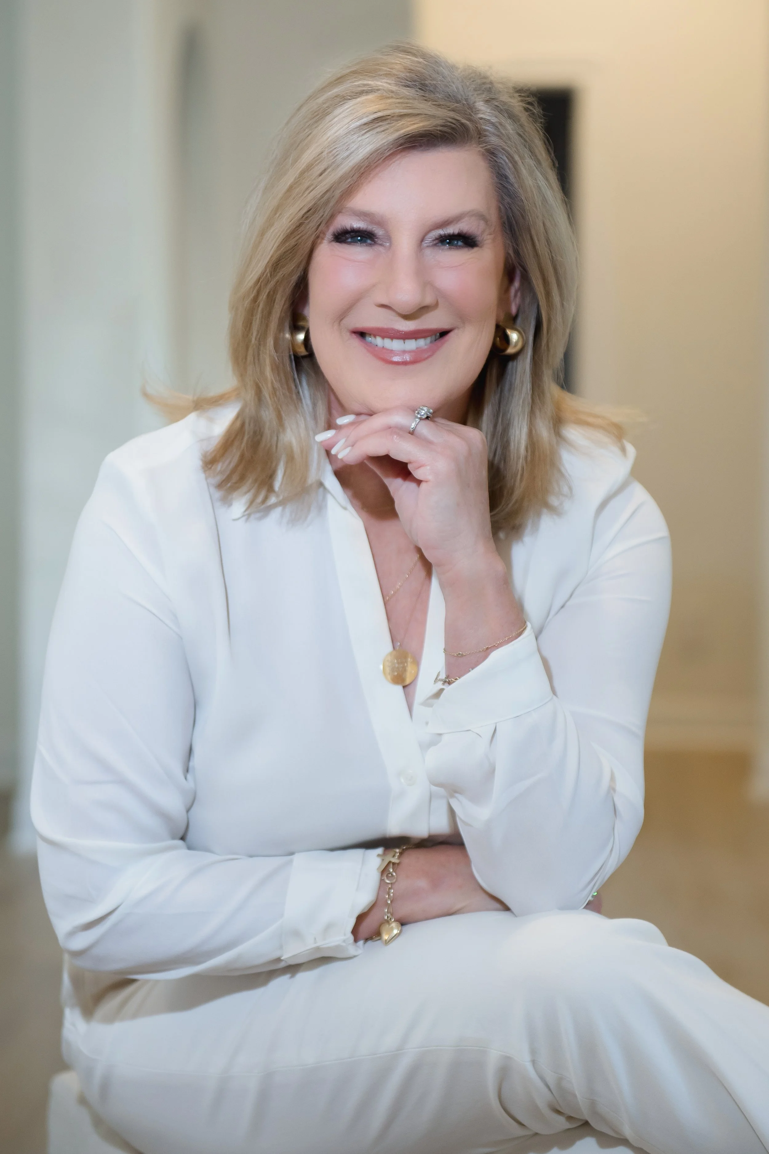 A smiling middle-aged woman with blonde hair, wearing a white blouse, gold jewelry, and earrings, sitting indoors with her chin resting on her hand.