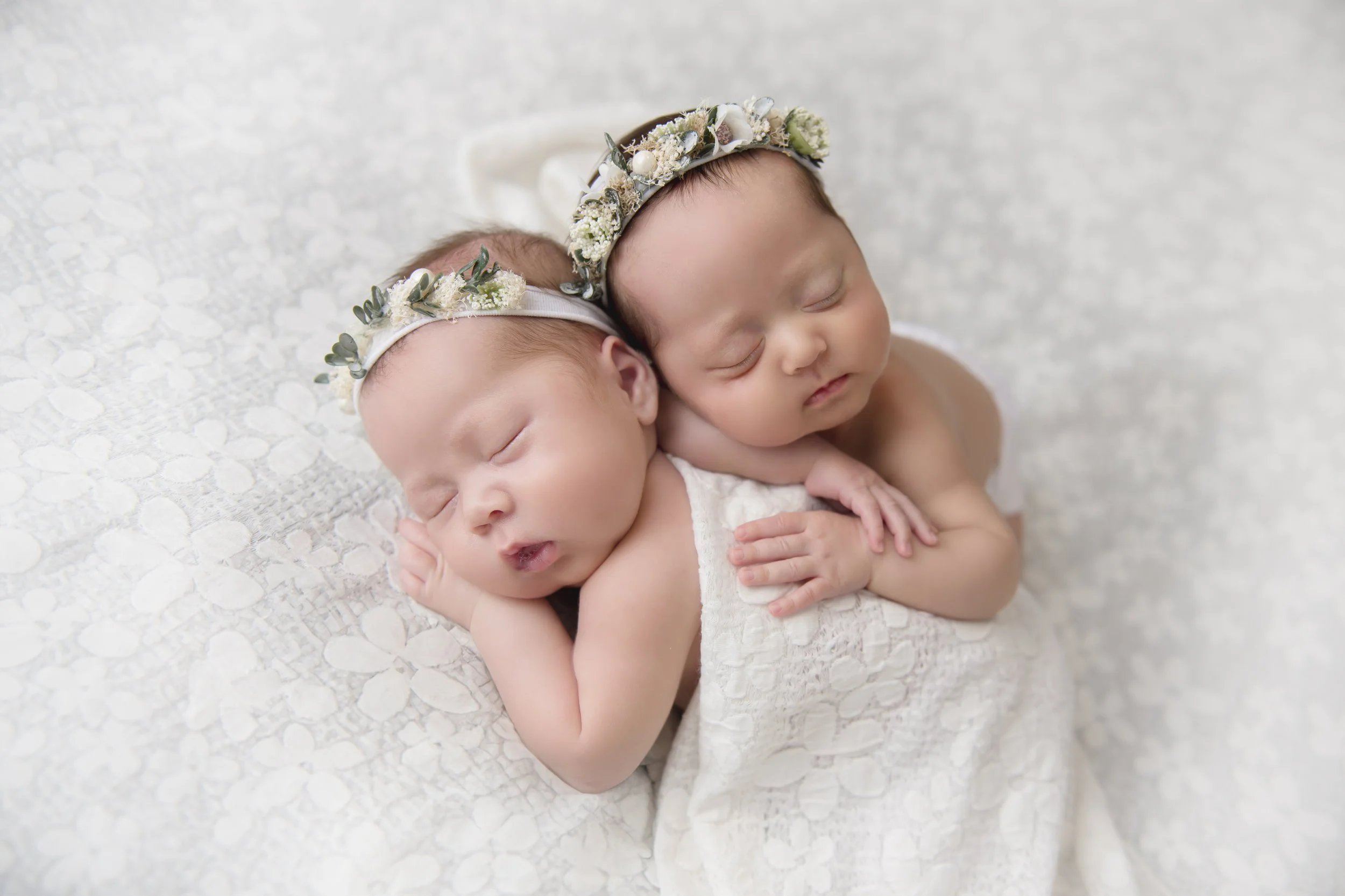 Heirloom twin newborn portrait of two sleeping babies wearing delicate floral headbands, posed closely together on a soft white blanket.