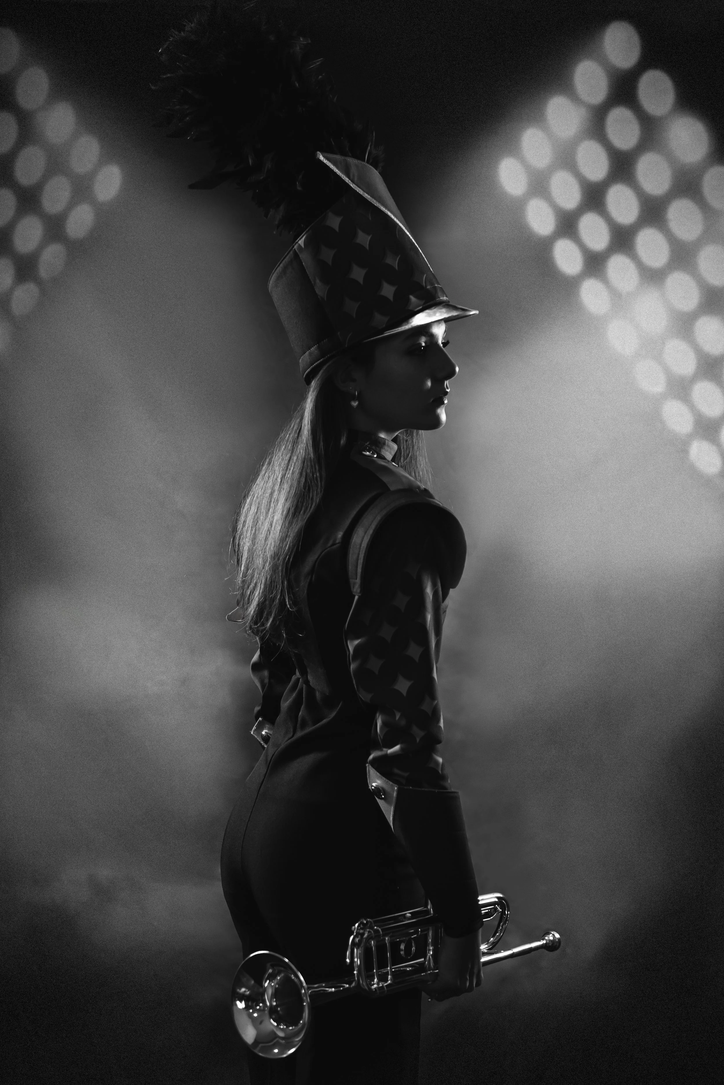 Black and white photo of a woman in a marching band uniform with a tall, decorated hat, holding a trumpet, standing in profile with stage lighting in the background.