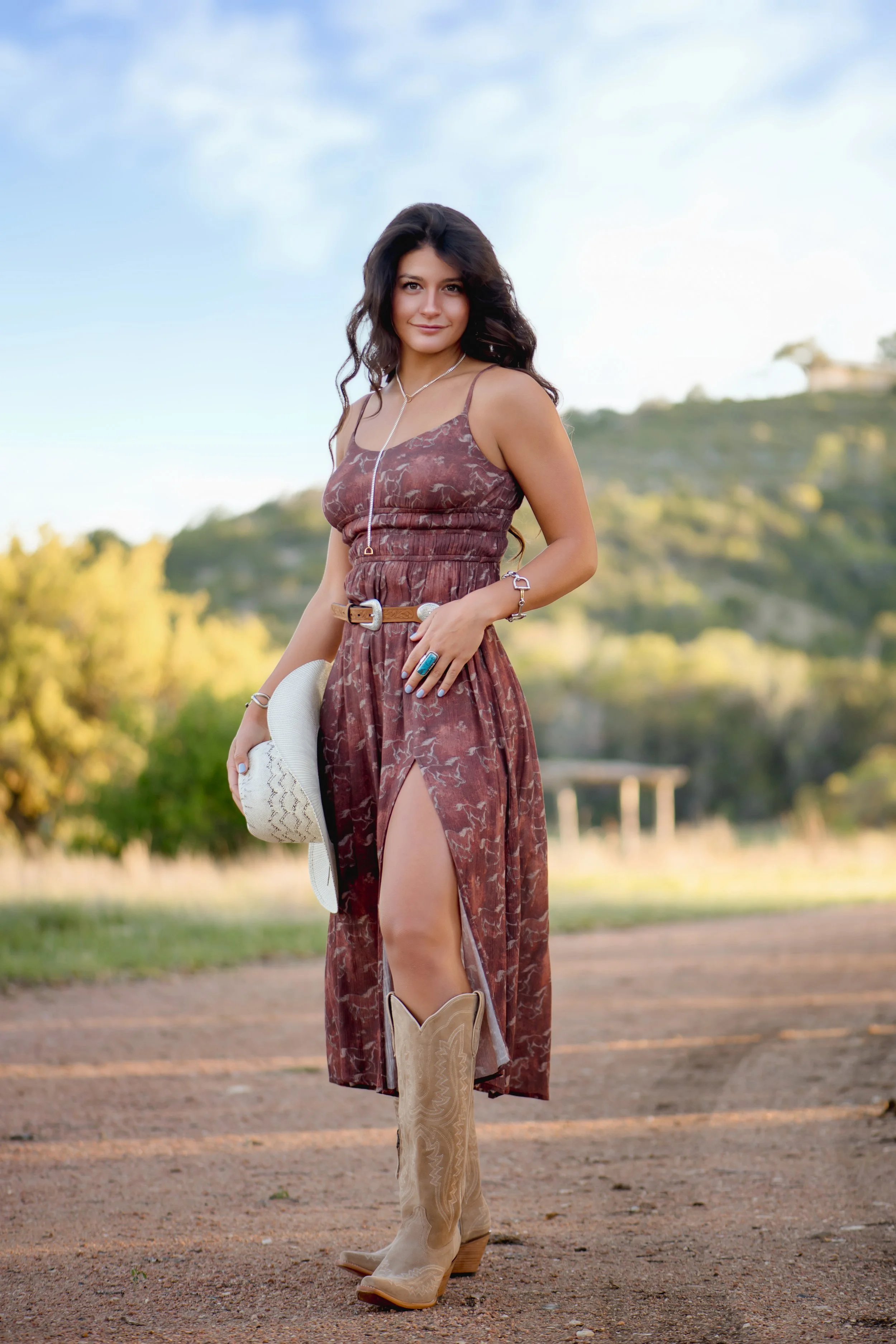 Young woman standing outdoors in a countryside setting, wearing a brown patterned dress with a high slit, cowboy boots, holding a white hat, and smiling at the camera.