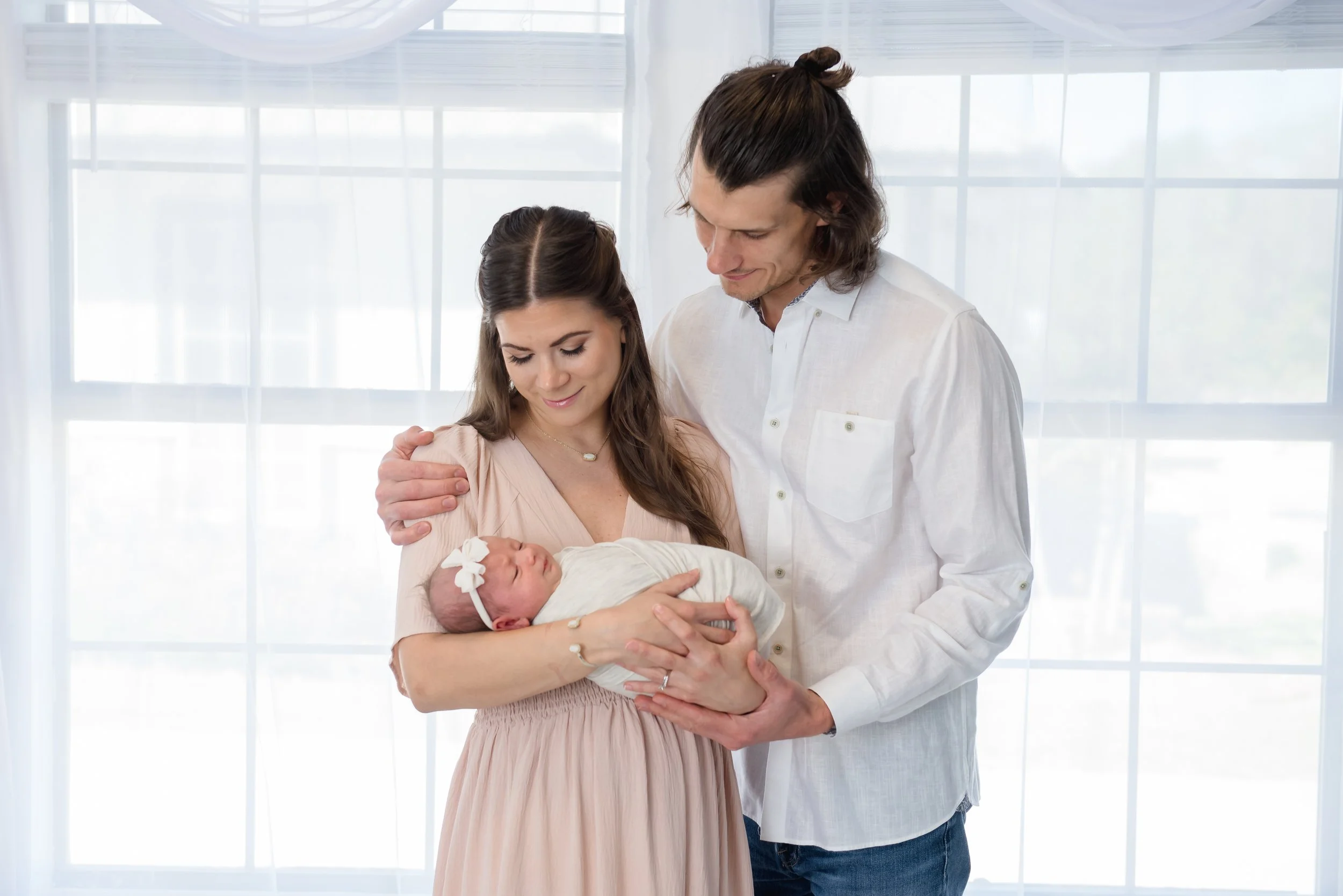 A young couple holding a newborn baby girl, standing in front of large windows with white curtains, in a bright room.