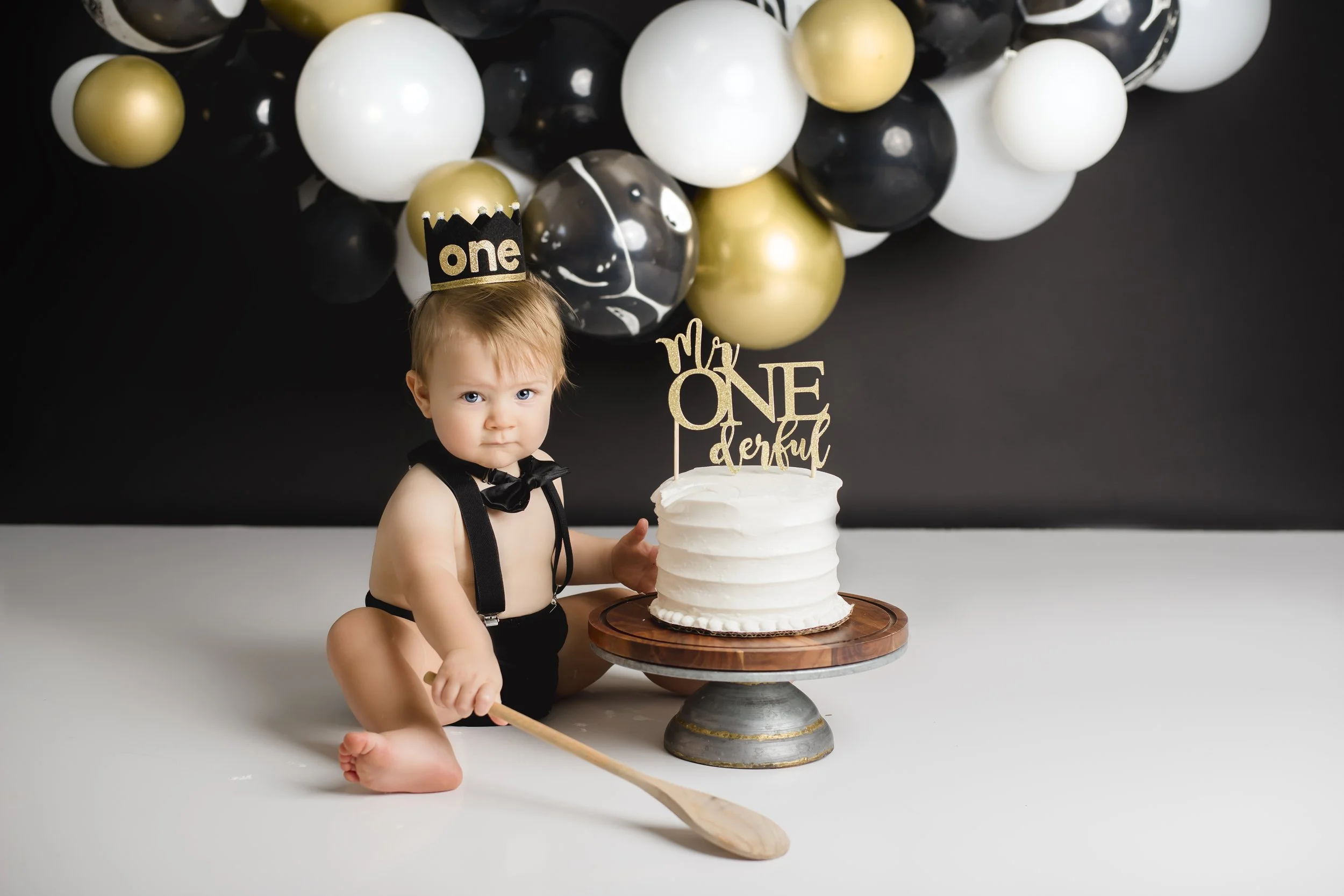 A toddler with a serious expression, sitting on a white floor, dressed in black suspenders and a bow tie, next to a white frosted cake on a stand. The cake has a topper that reads 'My One Derful,' and the toddler wears a crown that says 'one.' There 