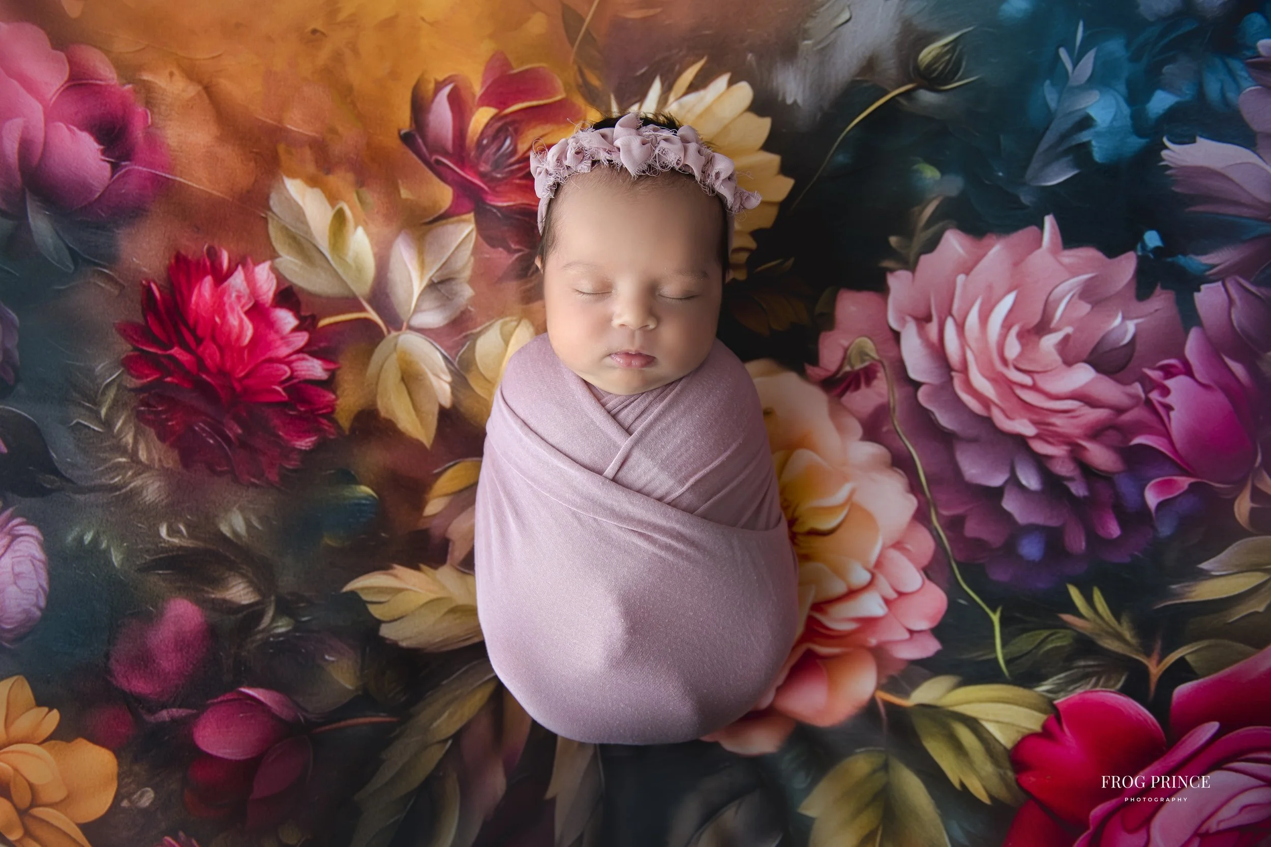A newborn baby swaddled in a pink blanket and wearing a pink headband, sleeping on a colorful floral backdrop with large pink and purple flowers.