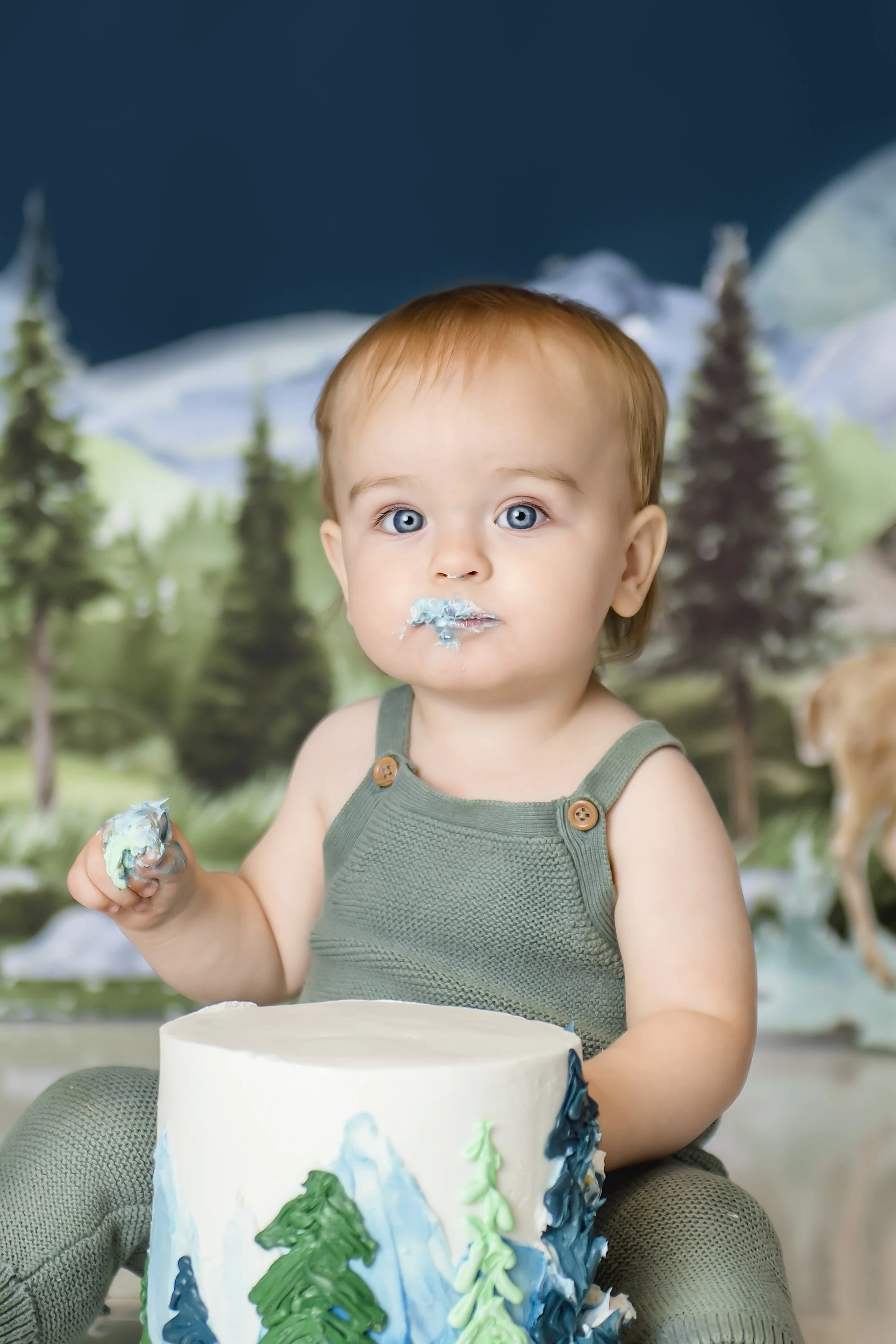 A young child with blue eyes and light brown hair is sitting in front of a cake with a forest mountain scene. The child has blue frosting around their mouth and is holding a piece of cake. The background features painted trees and mountains, creating