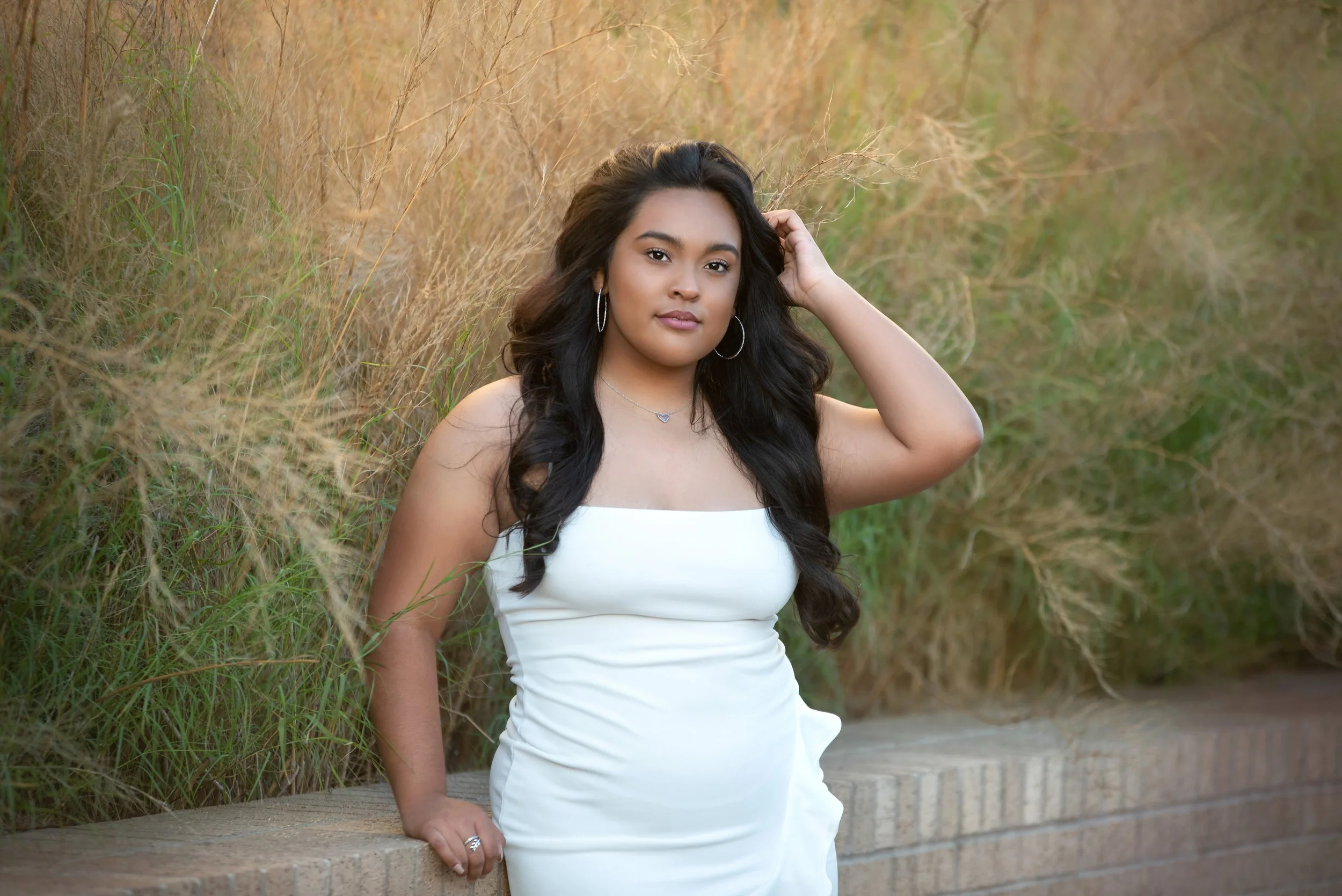 Young woman with long black hair in a white dress standing outdoors near grass and a brick wall.
