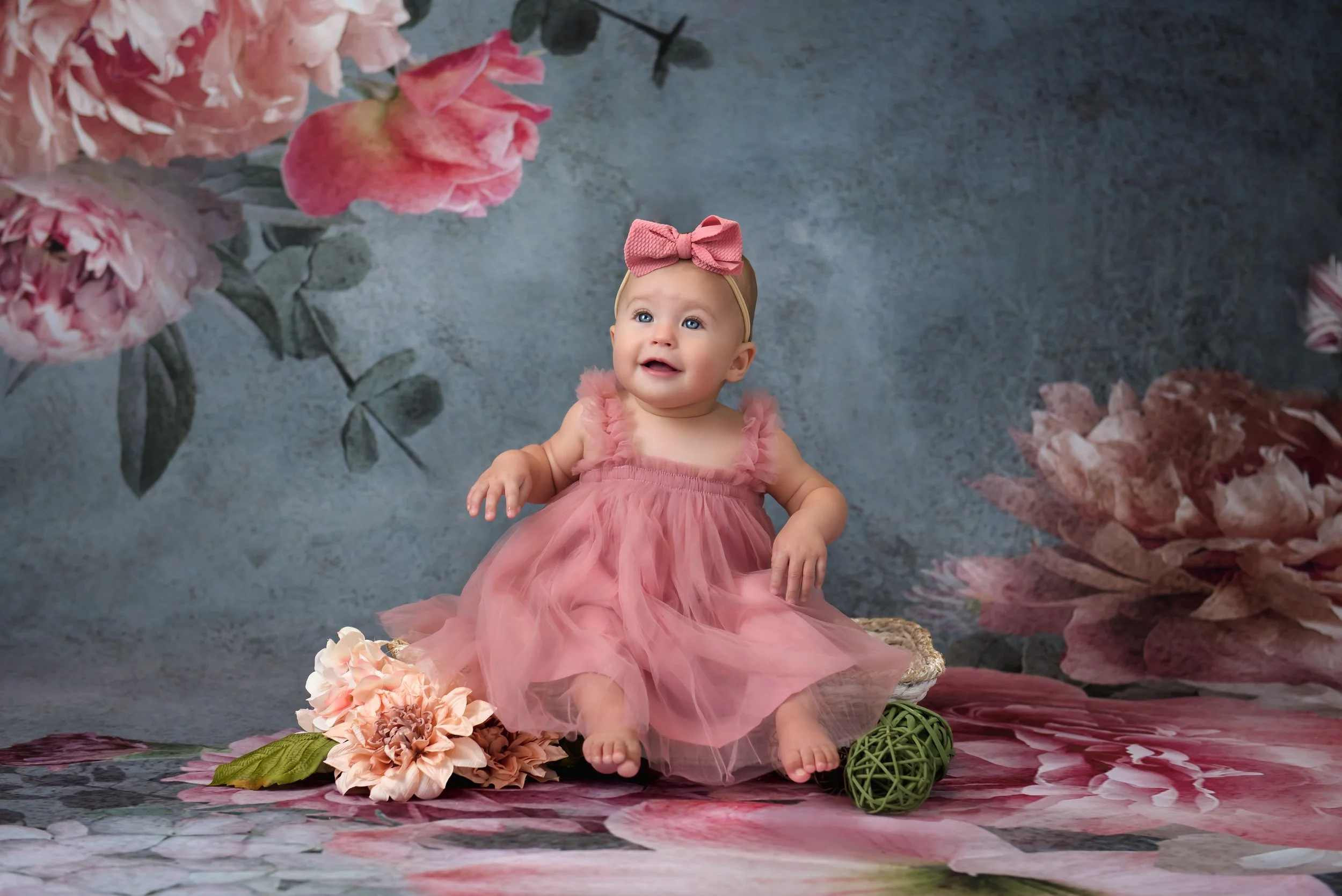 A baby girl in a pink dress with ruffled straps and a large pink bow headband, sitting on a floral fabric with a bouquet of flowers and a decorative ball, in front of a backdrop featuring large pink flowers and green leaves.