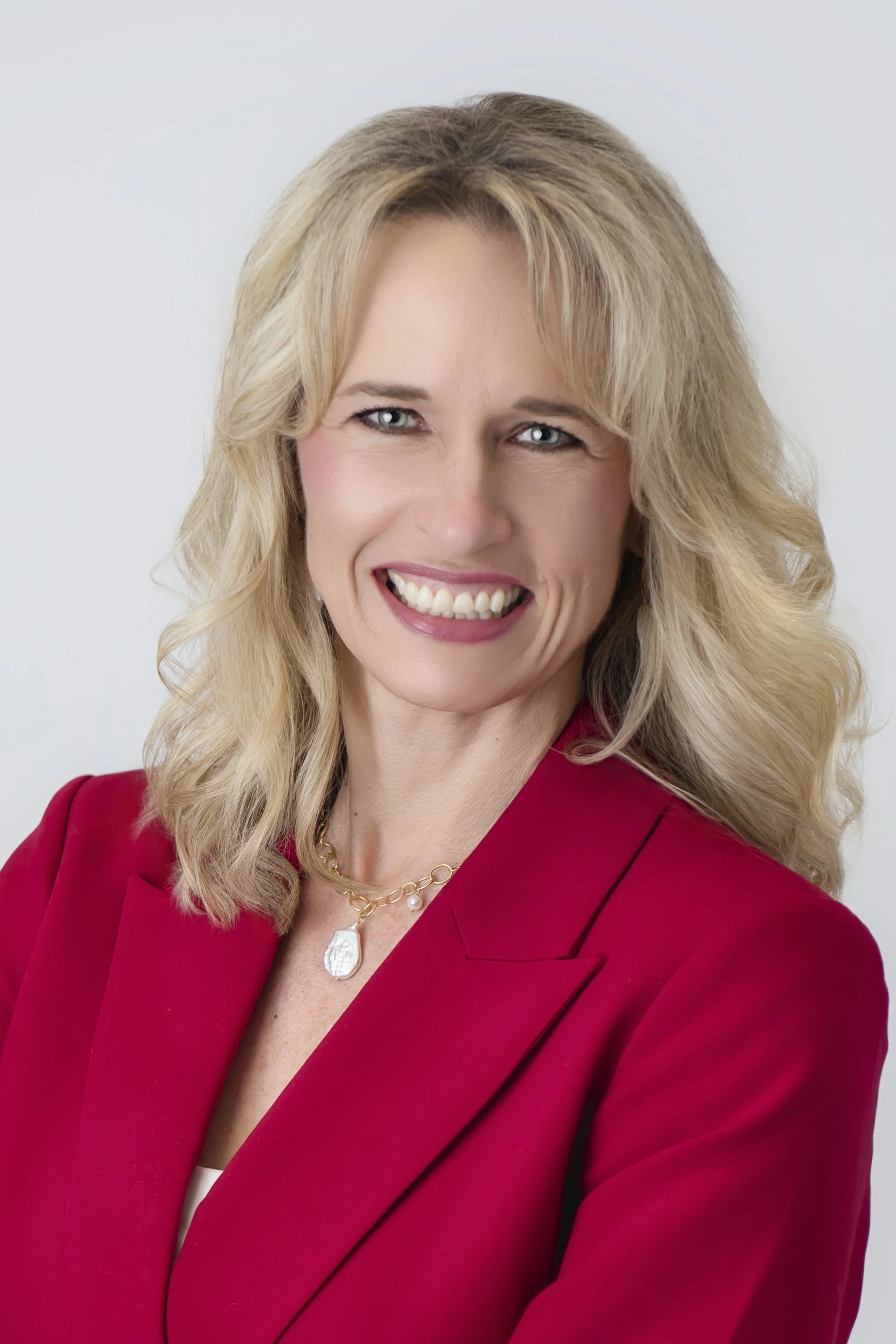 A woman with blonde wavy hair smiling, wearing a red blazer and a gold necklace with a large pendant, against a light gray background.