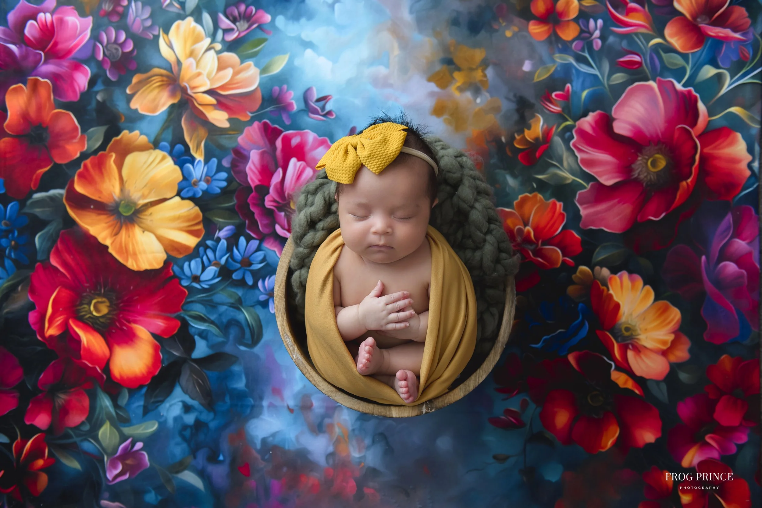 A sleeping baby with a yellow headband and matching cloth wrapped around the body, lying in a round wooden bowl on a vibrant, colorful floral backdrop.