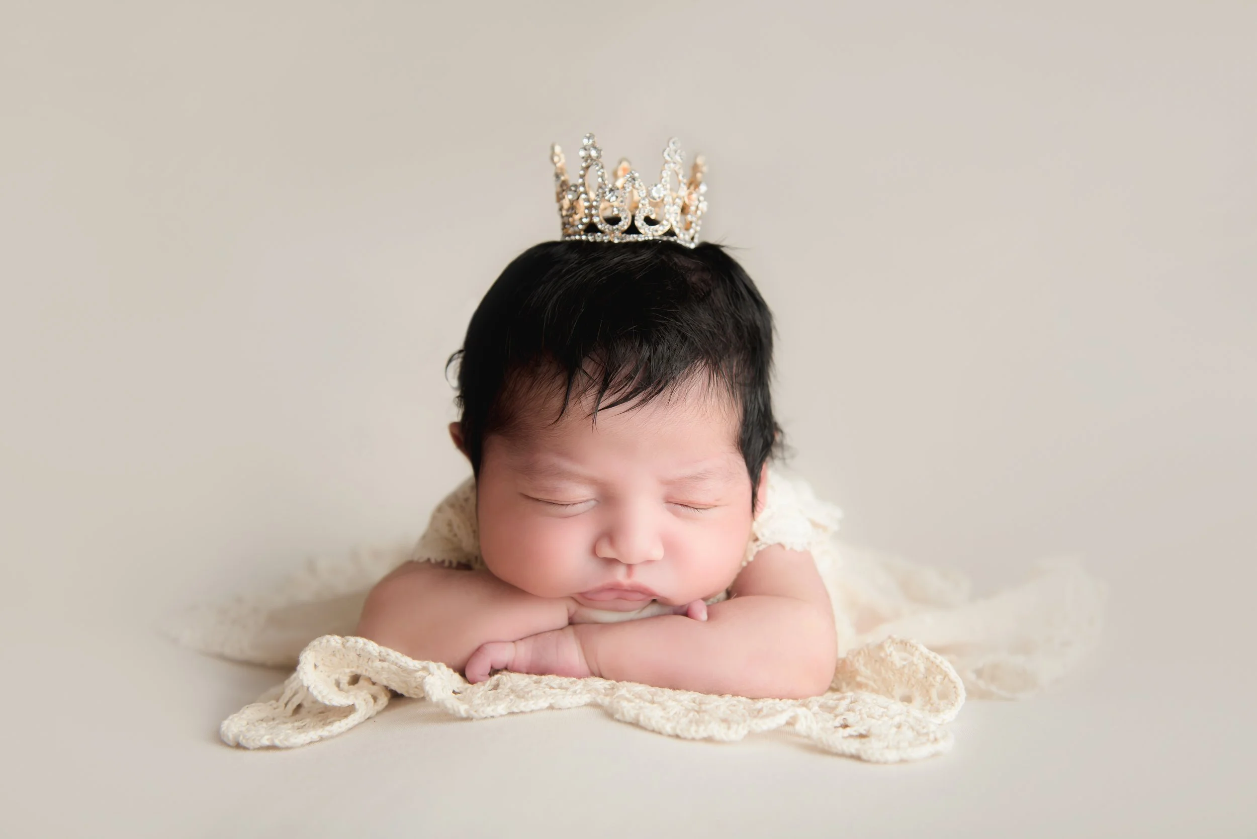 Luxury newborn portrait of a sleeping baby wearing a pearl and rhinestone crown, resting on a cream heirloom blanket.