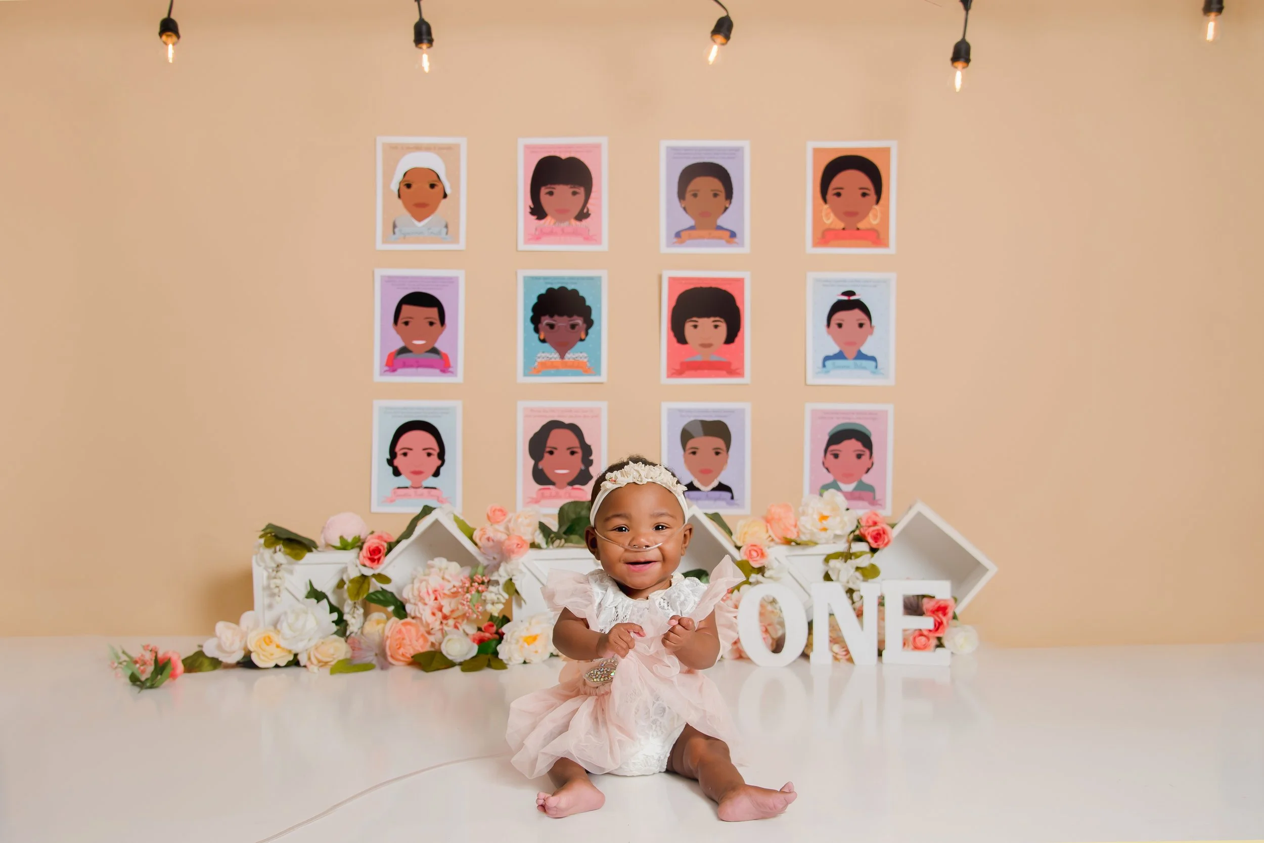 A baby girl celebrating her first birthday, sitting on a white surface in front of a floral backdrop with large white letters spelling "ONE" and wall art of illustrated portraits of diverse women.
