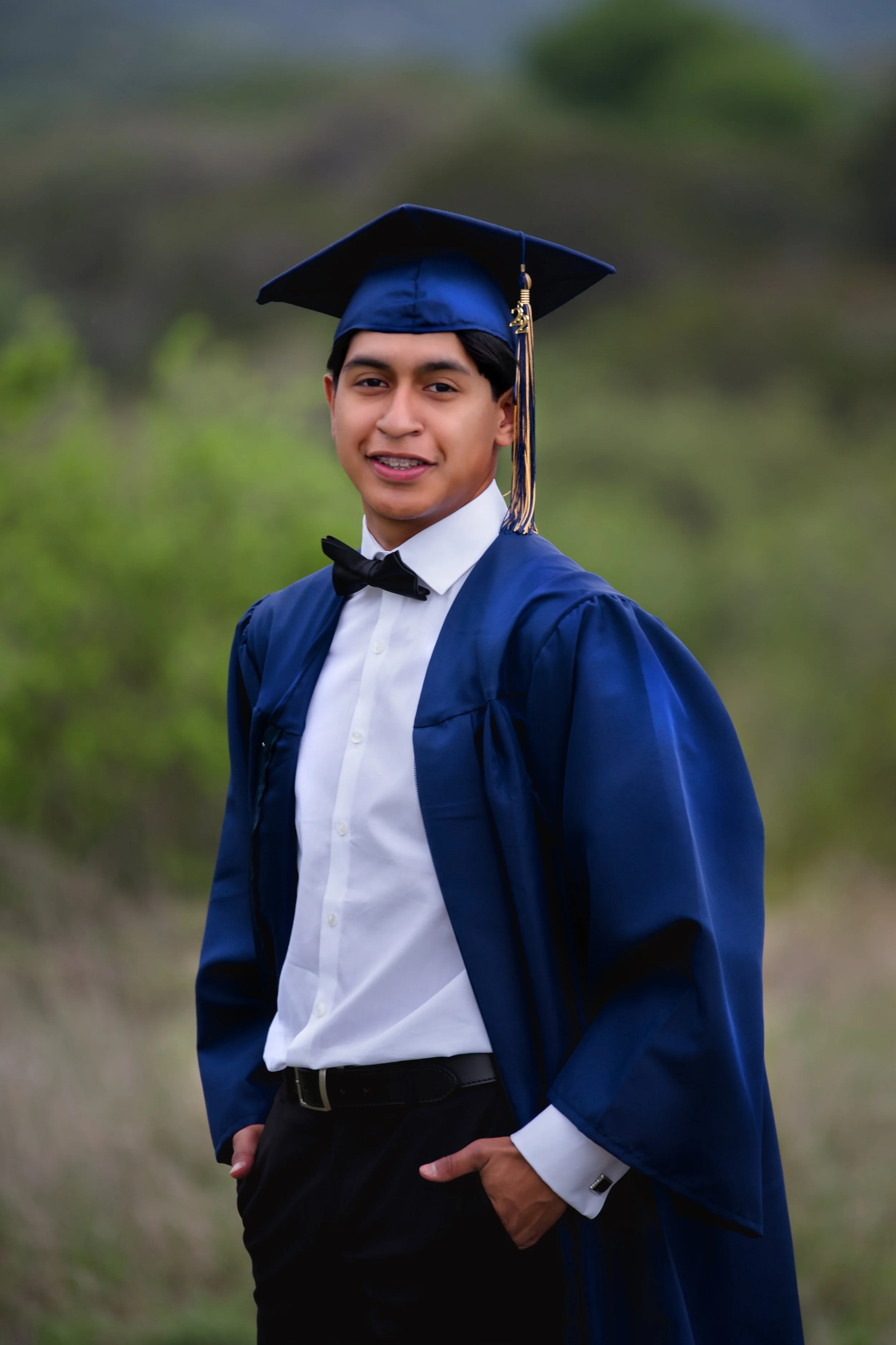 Young man in a graduation cap and gown smiling outdoors, with trees and greenery in the background.