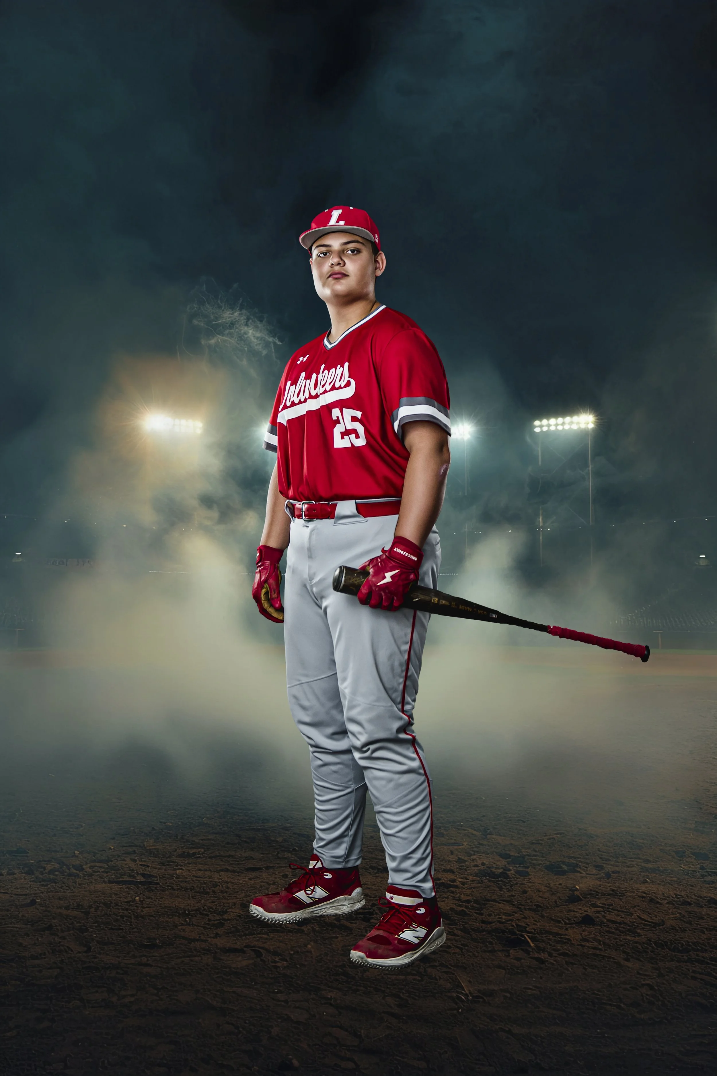 A young male baseball player dressed in a red jersey, gray pants, and a red cap, standing on a baseball field at night with stadium lights in the background, holding a baseball bat.