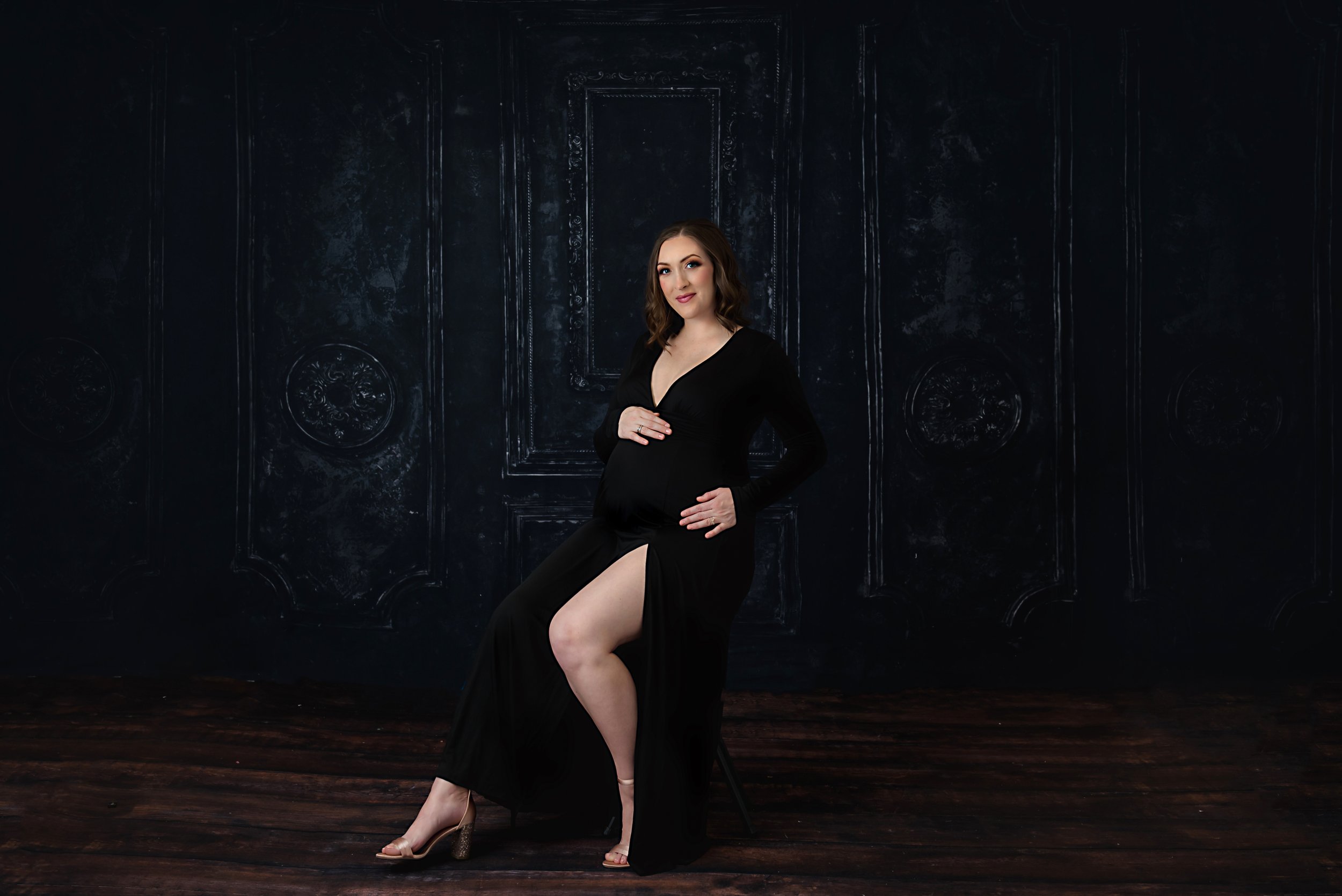 Editorial maternity portrait of a pregnant mother in a black gown with high slit, seated in an ornate studio setting with dramatic lighting.