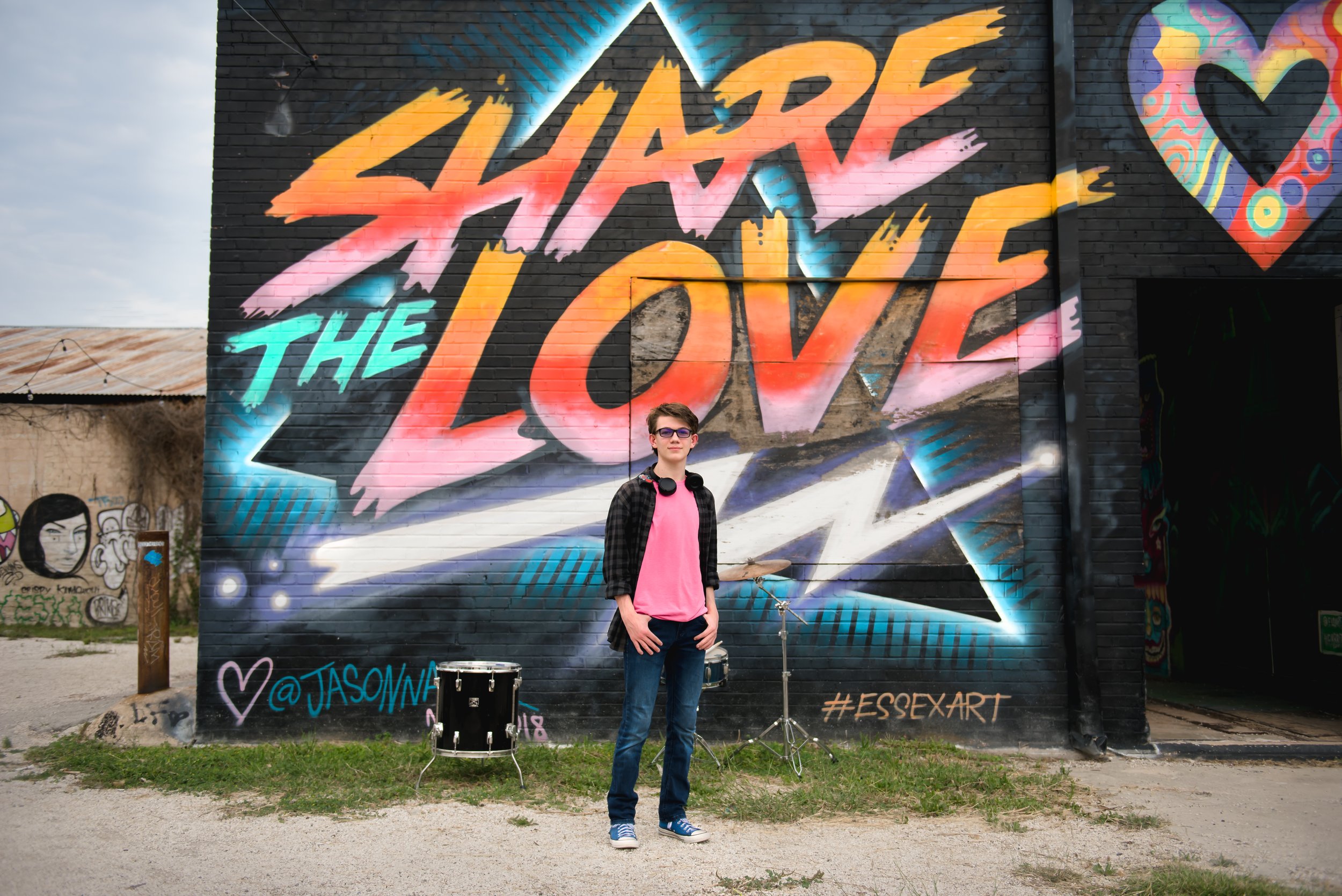 A young person standing in front of a colorful graffiti mural that reads 'Share the Love' with a heart symbol and social media handle '@JASONNA'. The person is wearing glasses, a pink shirt, a black checkered jacket, and jeans, with a small drum set 