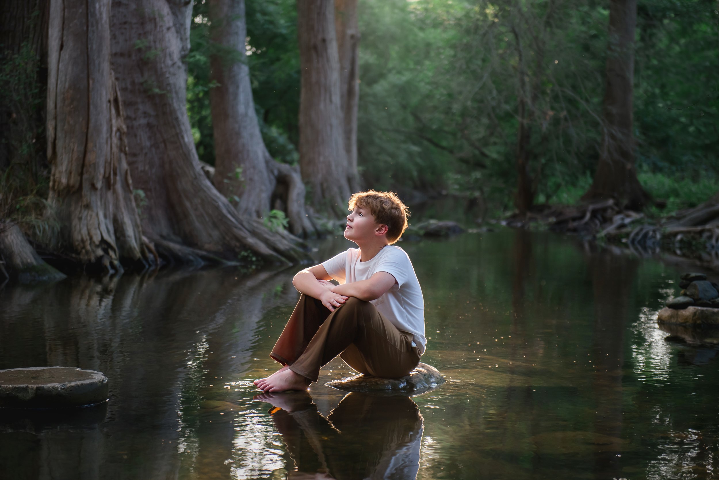 A boy sitting on a rock in a creek, surrounded by tall trees with green leaves, during daytime.