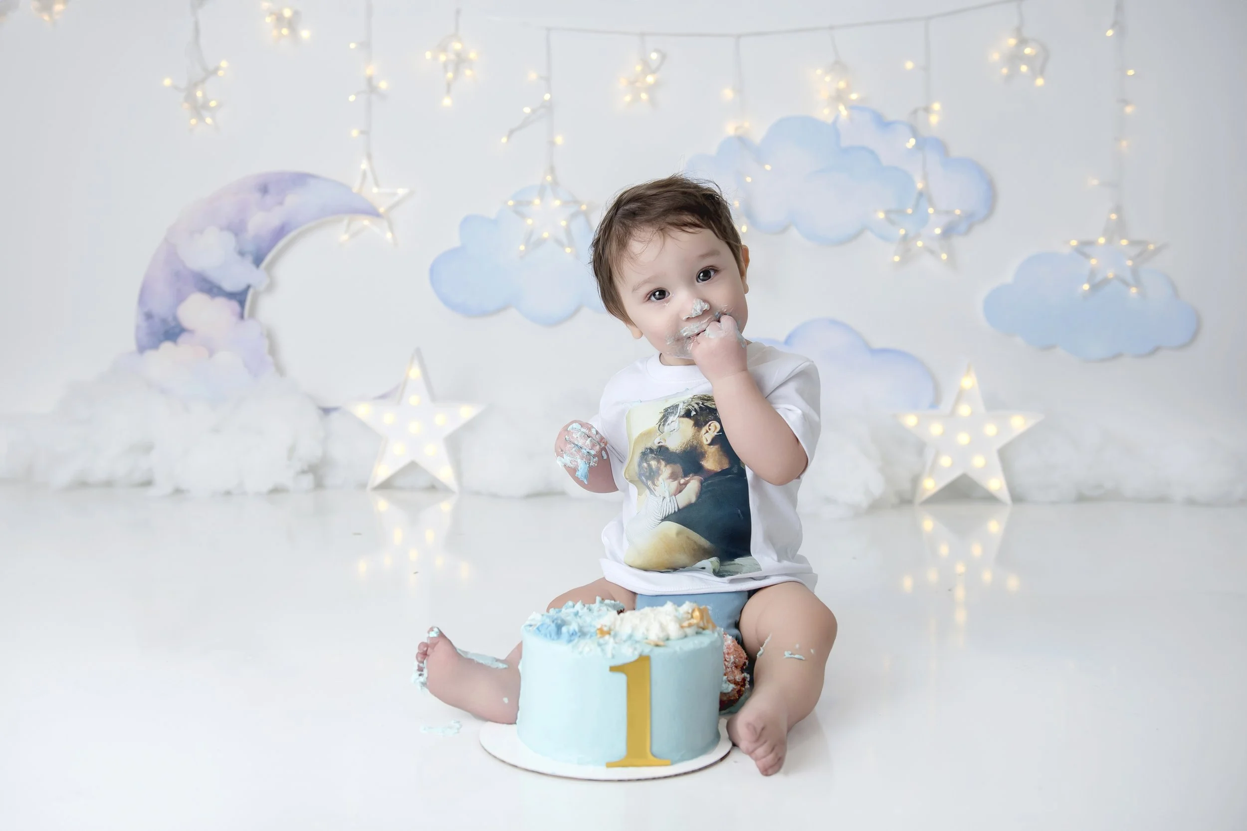 Cute toddler sitting on the floor celebrating first birthday with blue cake, wearing a white t-shirt with a graphic, surrounded by cloud and star decorations, some frosting on face and hands.