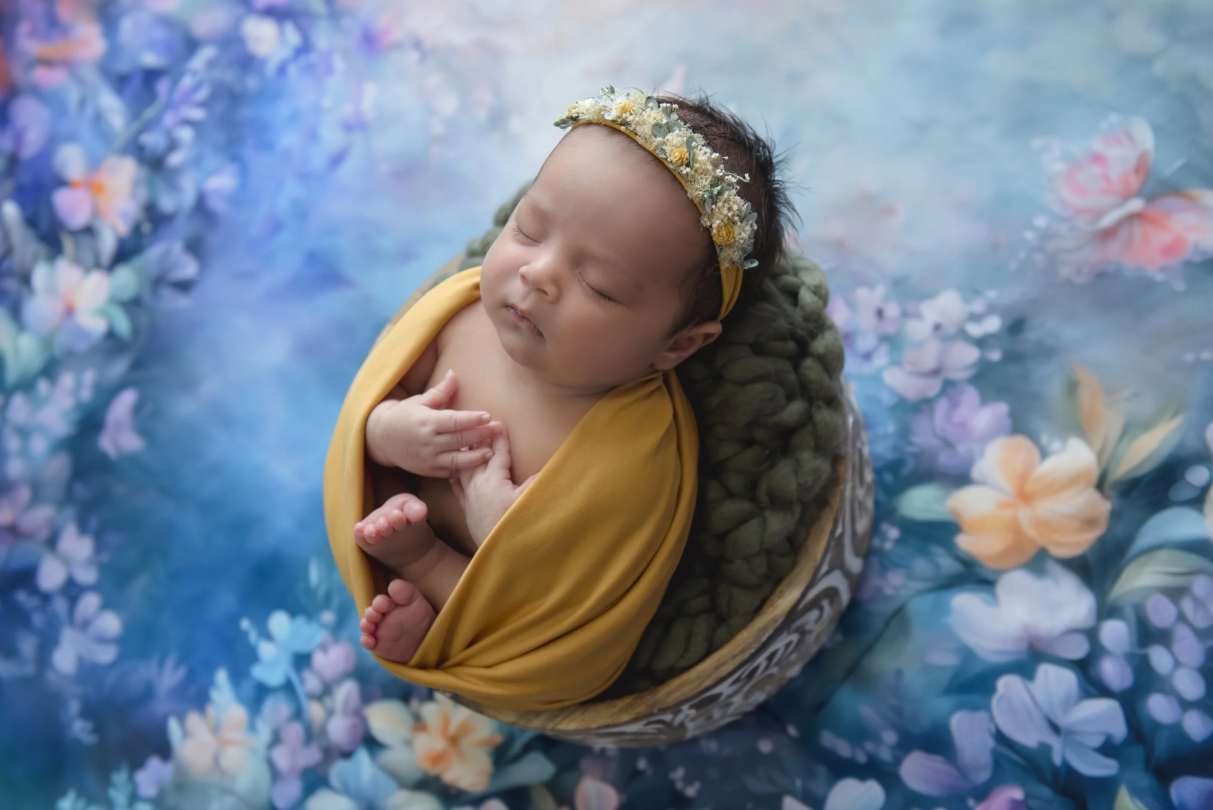 A sleeping newborn baby wrapped in a yellow blanket, lying in a bowl on a floral-themed background