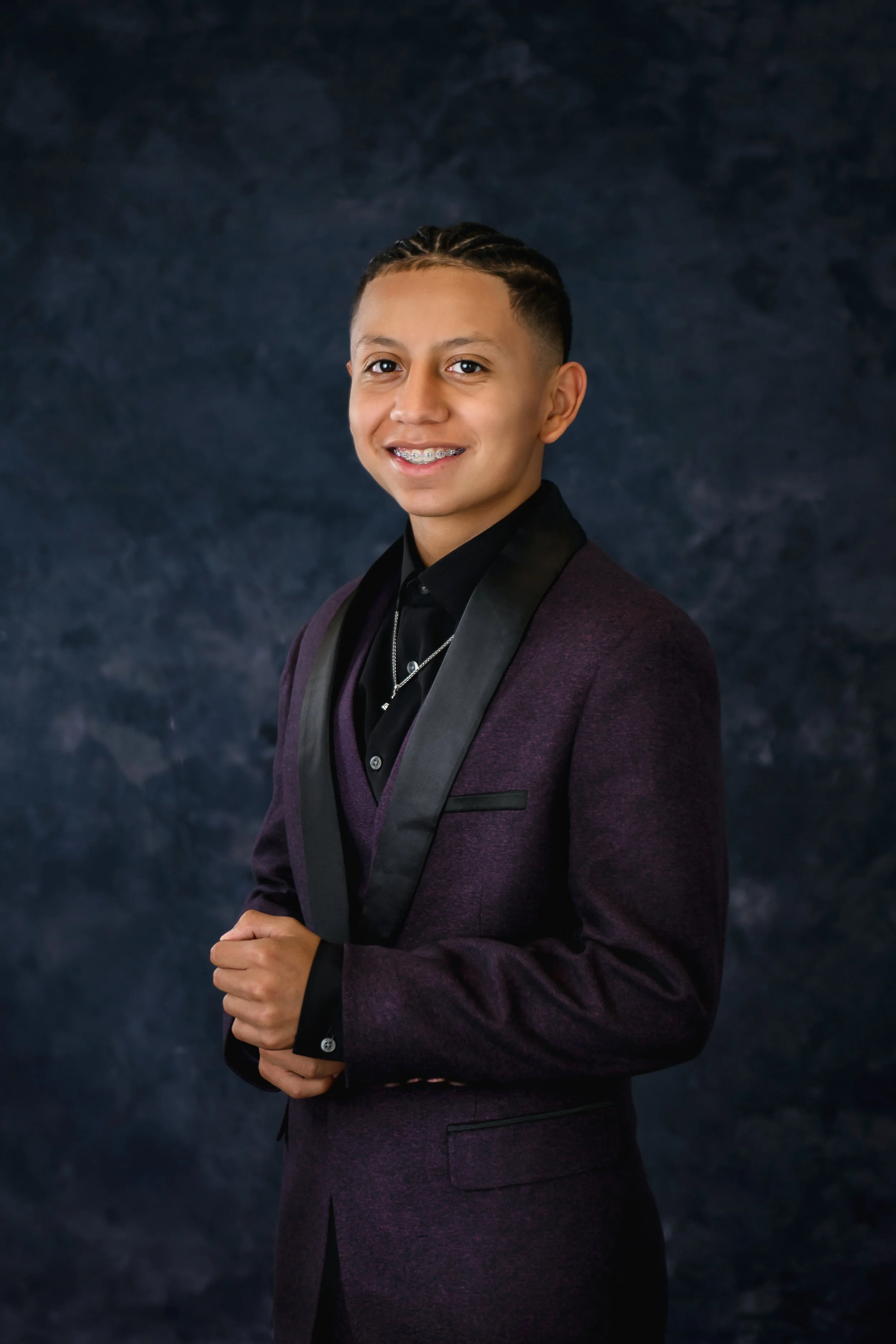 Young man with braces dressed in a dark purple tuxedo and black shirt, smiling at the camera against a dark textured background.