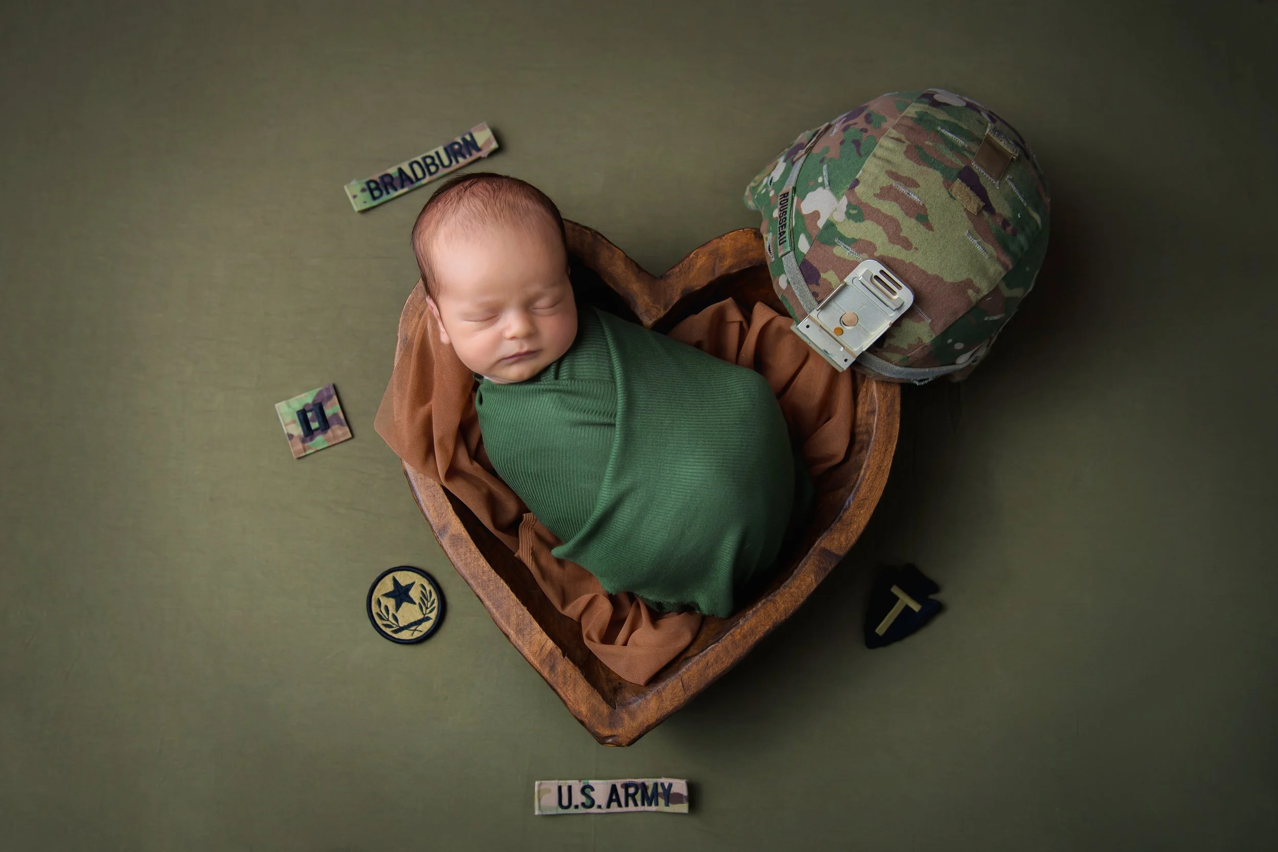 A sleeping baby wrapped in a green blanket, lying in a wooden heart-shaped box, with U.S. Army patches, a camouflage helmet, and a name tag around.