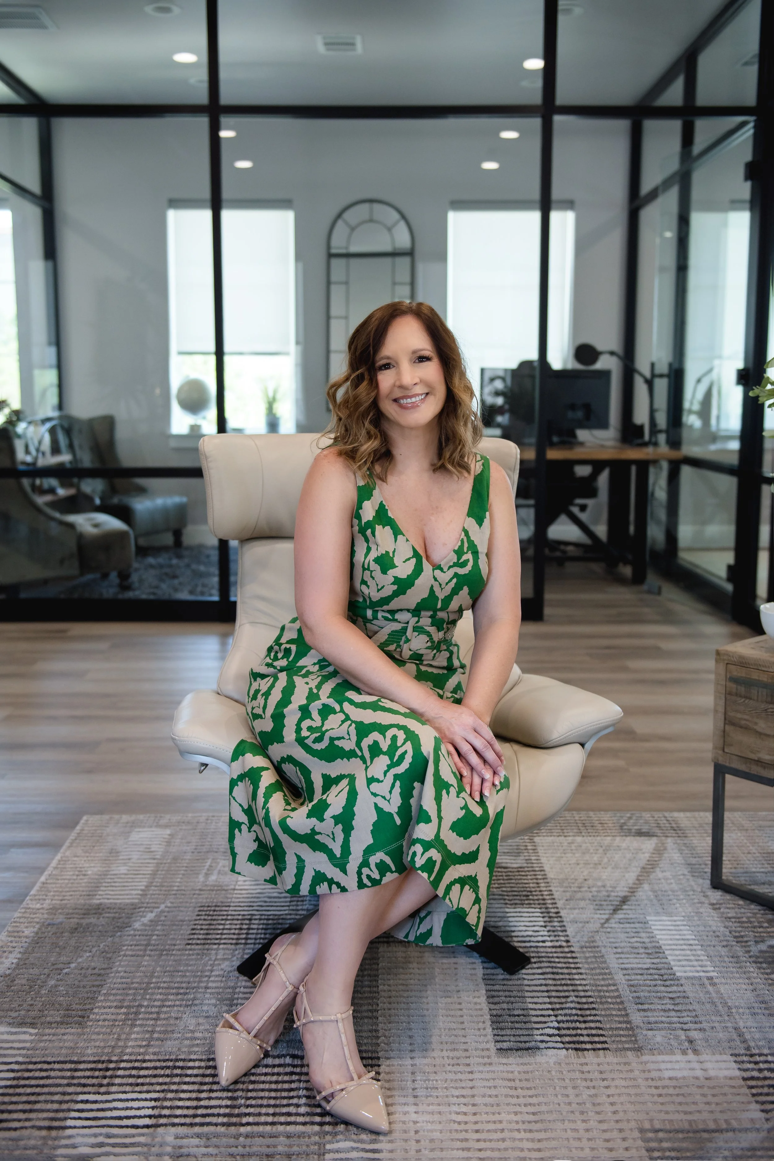 A woman with shoulder-length wavy brown hair, wearing a green and beige floral dress, smiling while sitting on a modern cream-colored chair in a bright, contemporary office space.