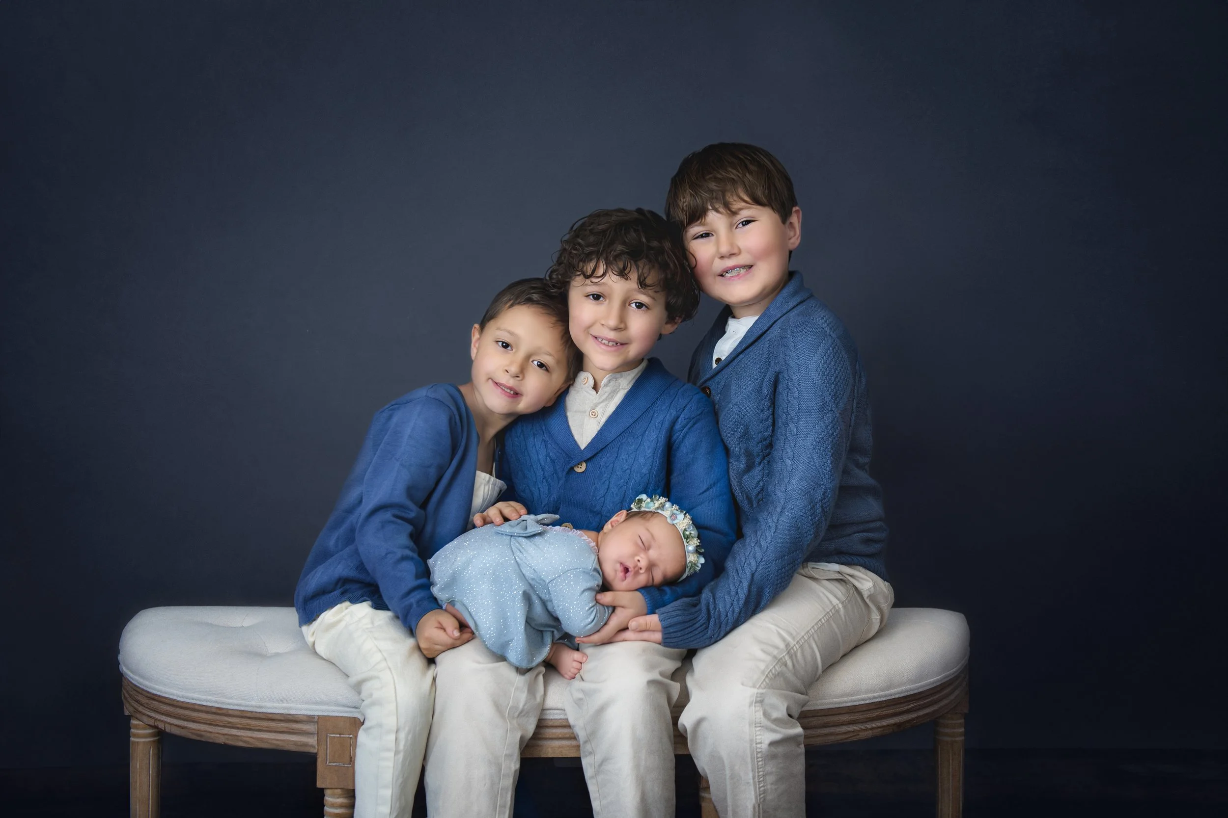 Four young boys sitting together on a cushioned bench against a dark background, with one of them being a sleeping baby girl dressed in blue with a floral headband.