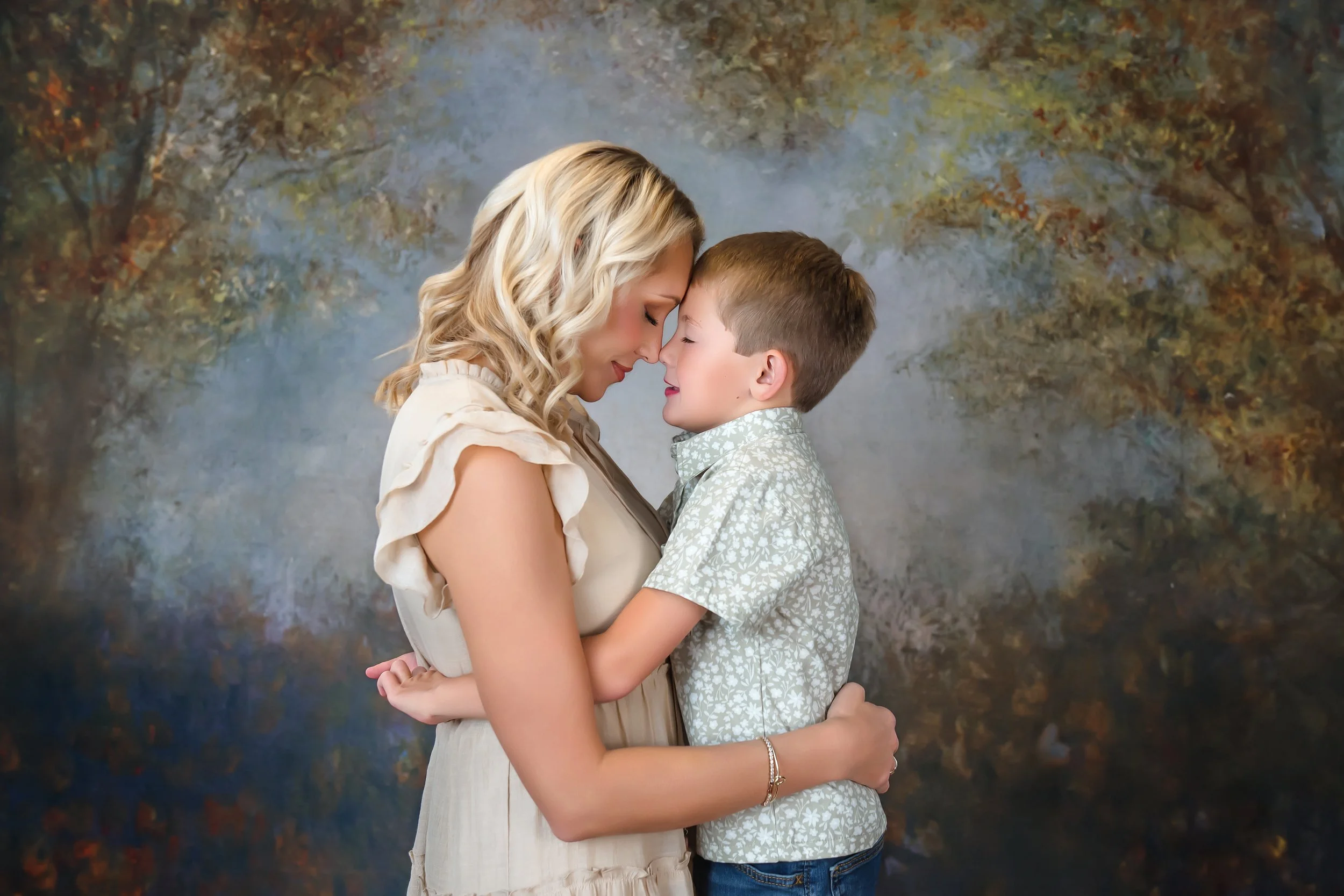 A woman and a young boy touching foreheads and smiling with eyes closed, standing closely together in front of a autumn landscape backdrop.