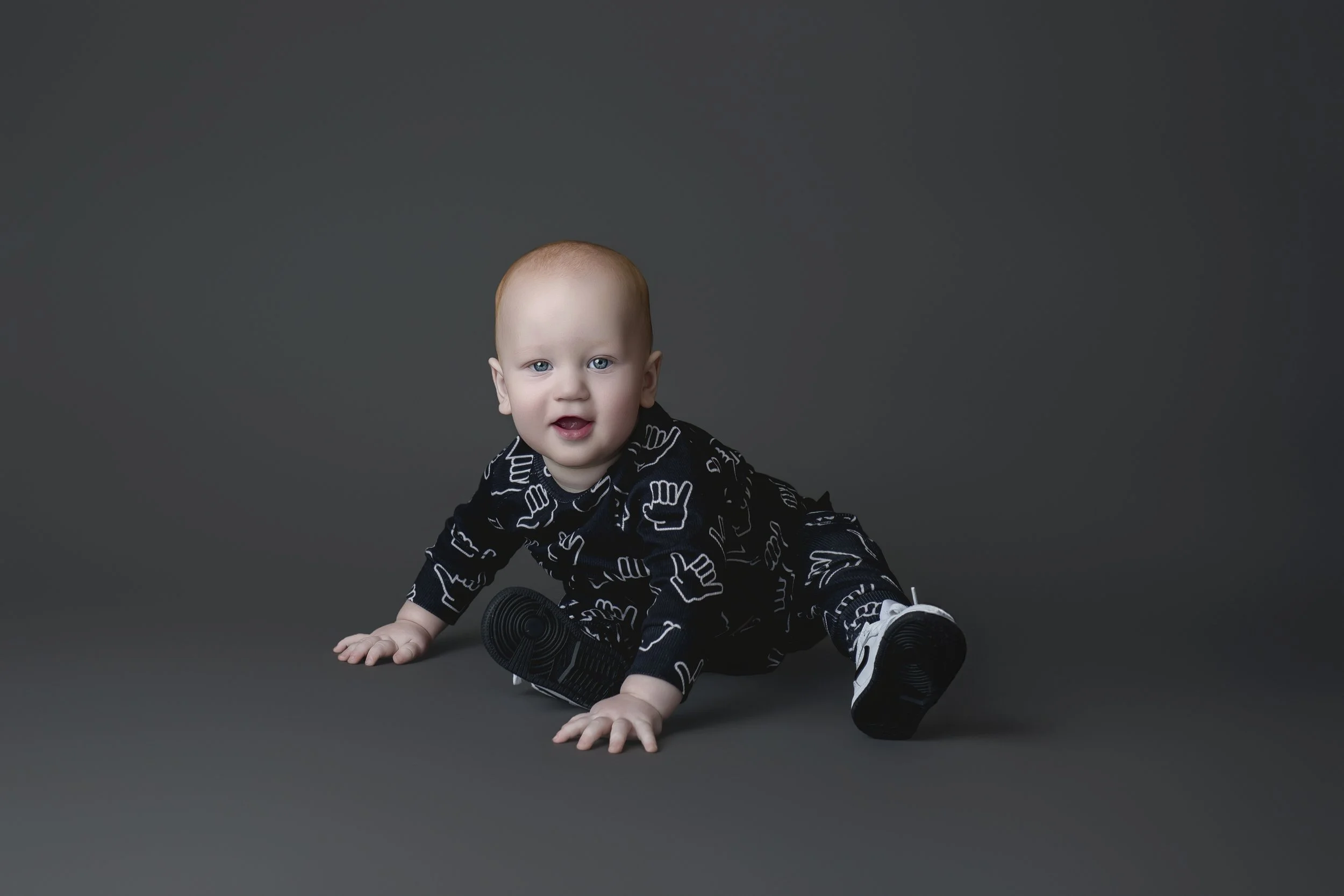 A baby with red hair and blue eyes in a black outfit with white hand signs, crawling on a gray background, wearing sneakers.