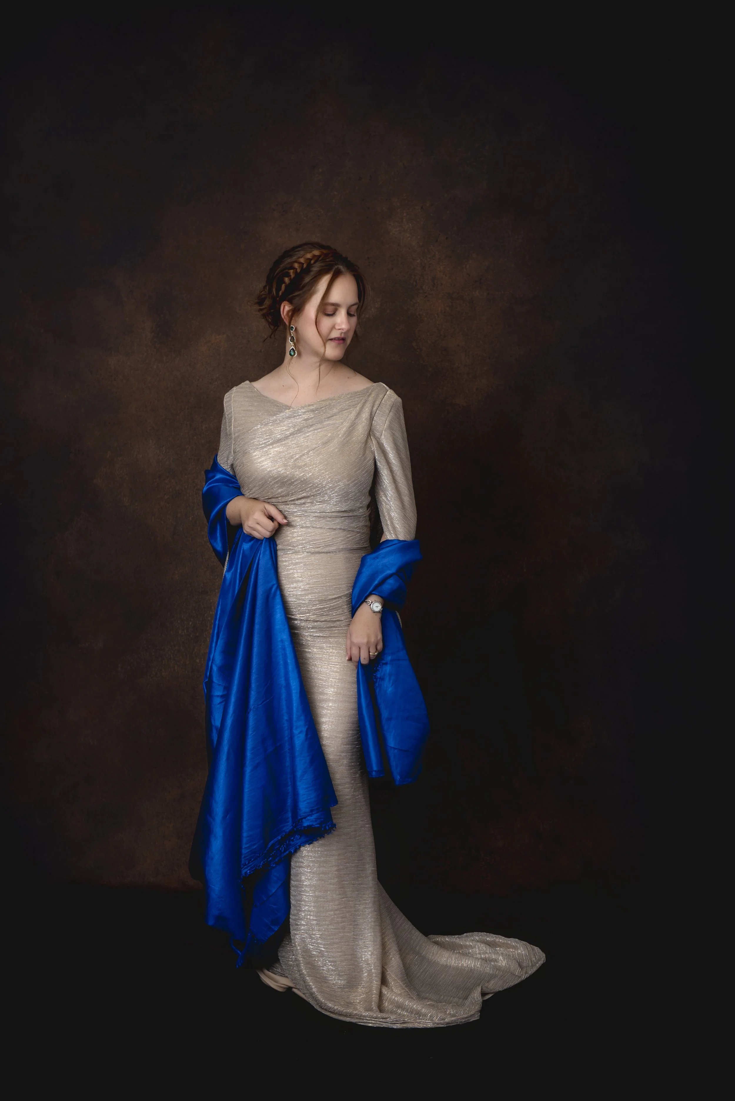 A woman in a gold dress with a blue shawl standing against a dark brown textured background, looking down.