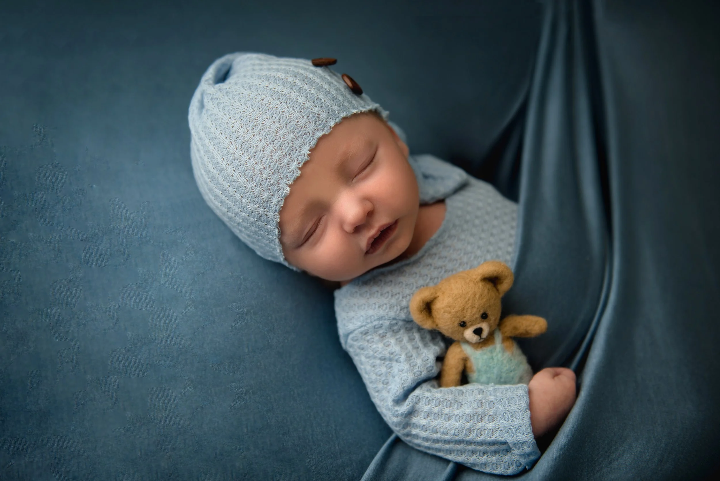 A sleeping baby wearing a light blue knit hat with brown buttons, cuddling a small teddy bear, lying on a blue surface.