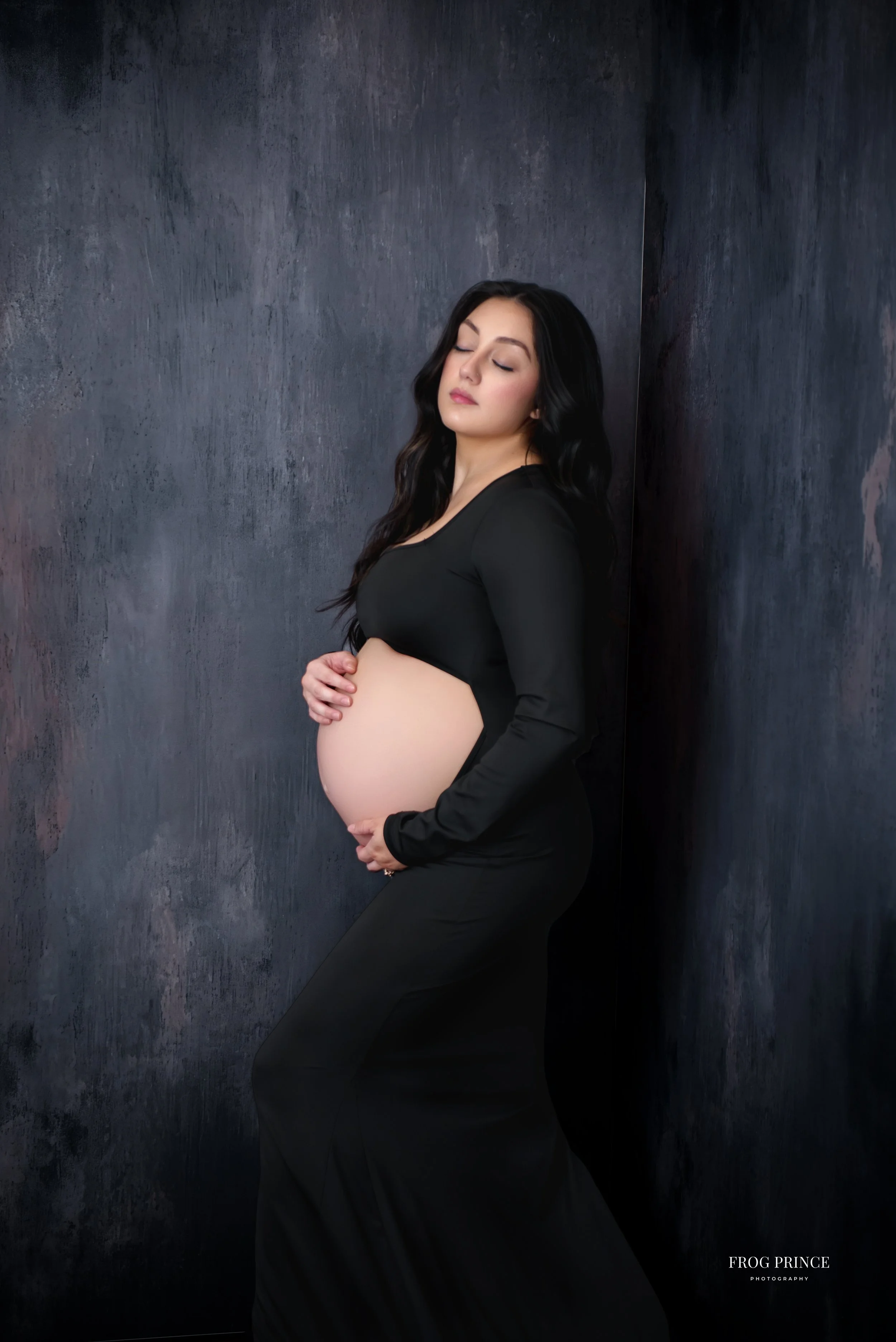 Editorial maternity portrait of a pregnant woman in a fitted black outfit, standing against a textured dark wall with eyes closed while gently cradling her baby bump in our Spring Branch studio.