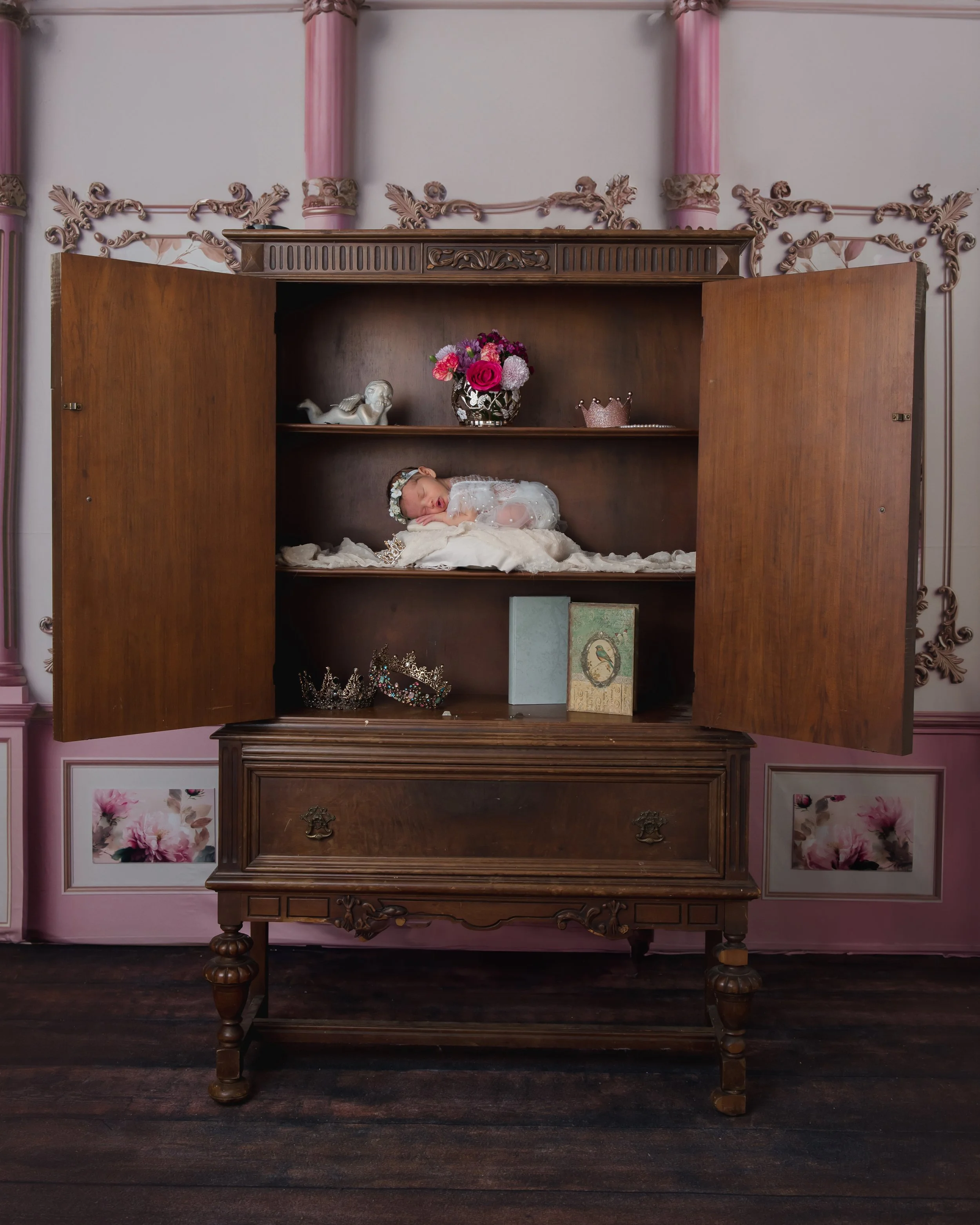 A baby girl dressed in white, sleeping on a blanket inside an open wooden cabinet with decorative objects and pink flowers above.